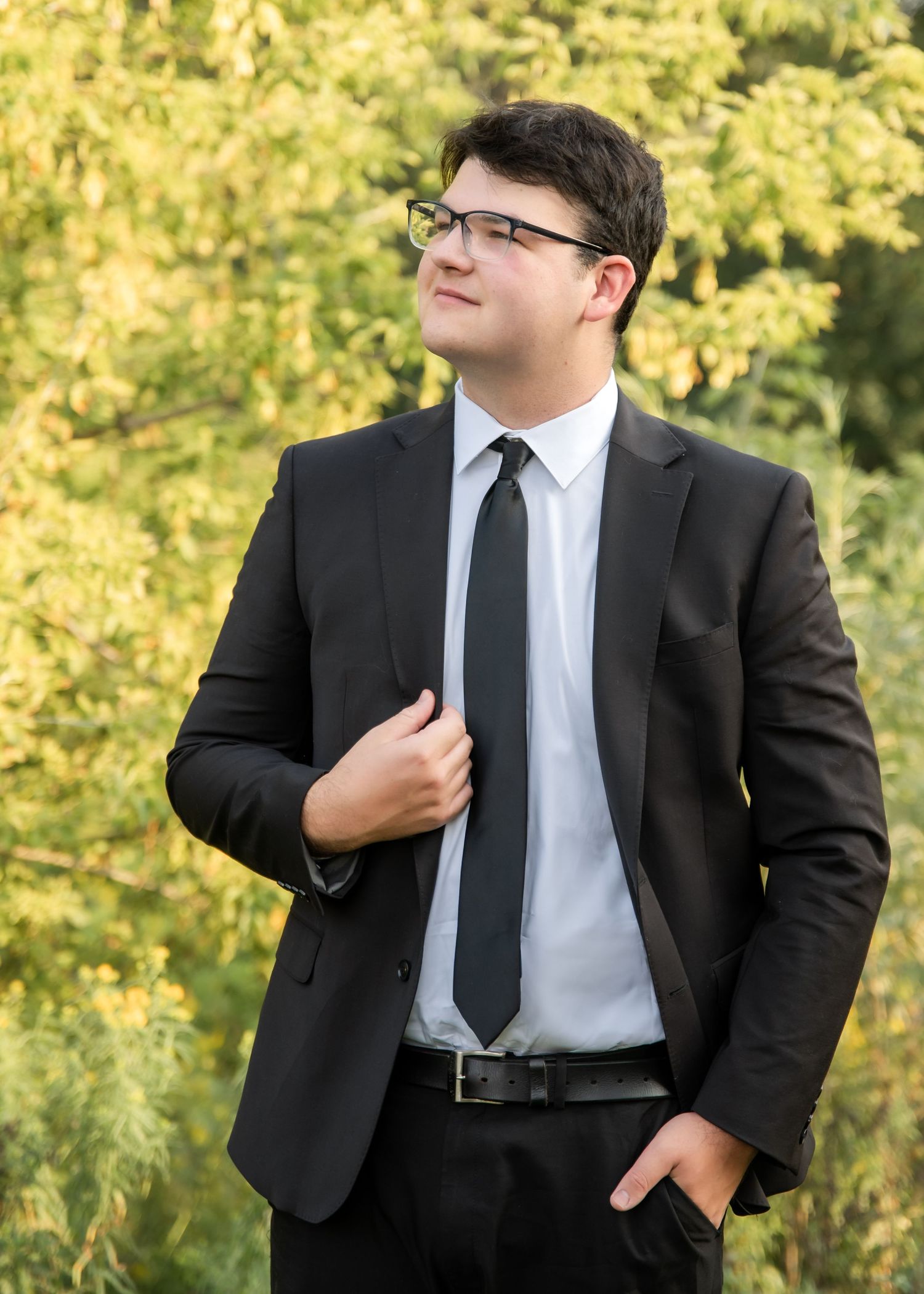 Professional figure wearing a black suit and tie adjusts his collar against a backdrop of autumn foliage.