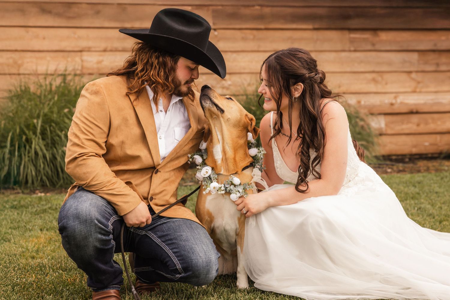 A couple in western-themed wedding attire sits on grass with their dog outside a rustic wooden wall.