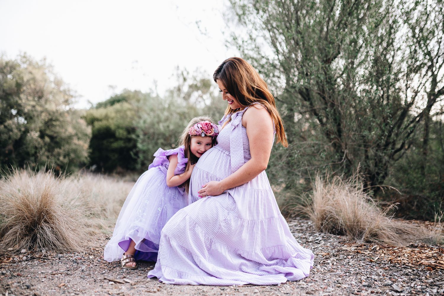 A tender moment between two figures in matching lavender dresses among tall grasses outdoors taken by Melbourne maternity photographer.