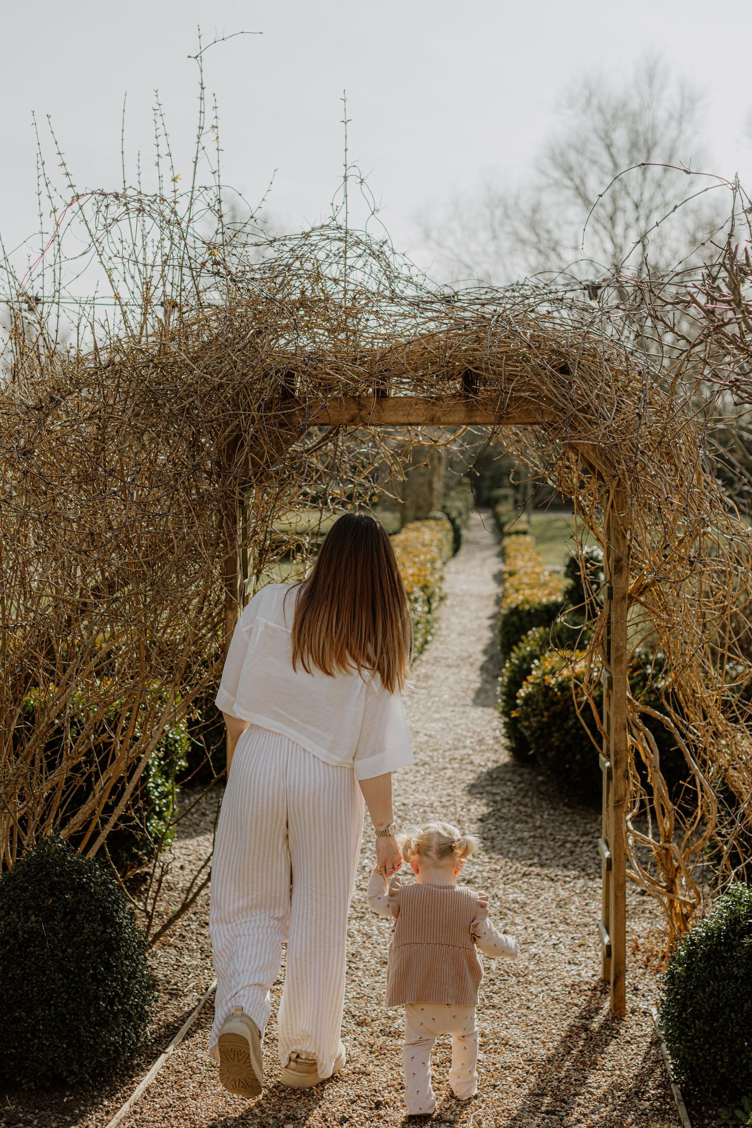 A parent and child walk down a garden path under a rustic wooden archway surrounded by bare vines.