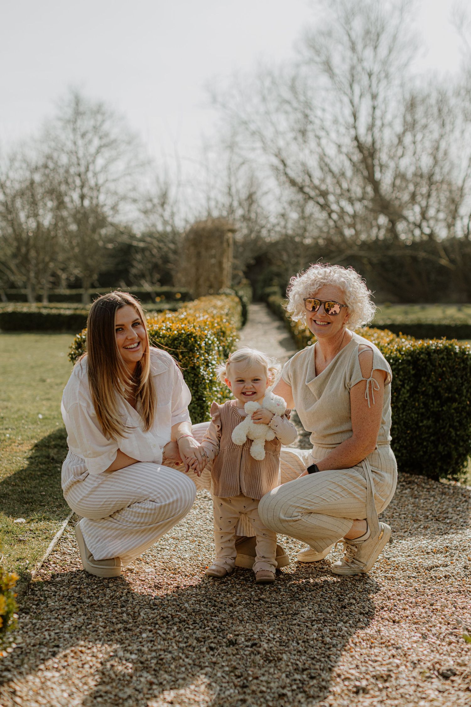Three generations pose together on a sunny day in a manicured garden setting.