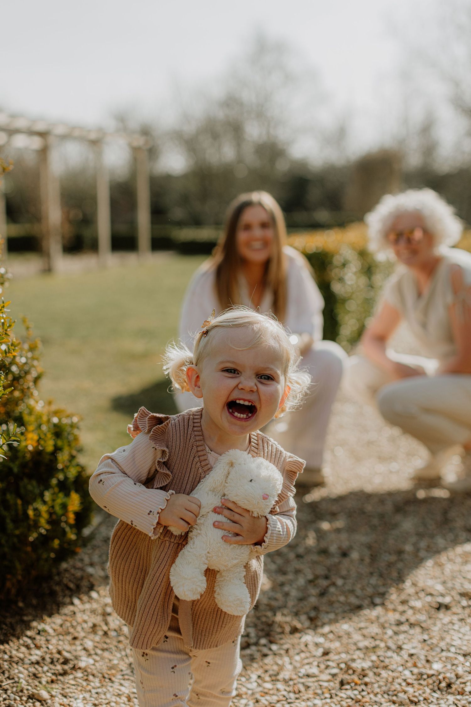 A joyful toddler laughs while holding a stuffed toy in a garden setting.