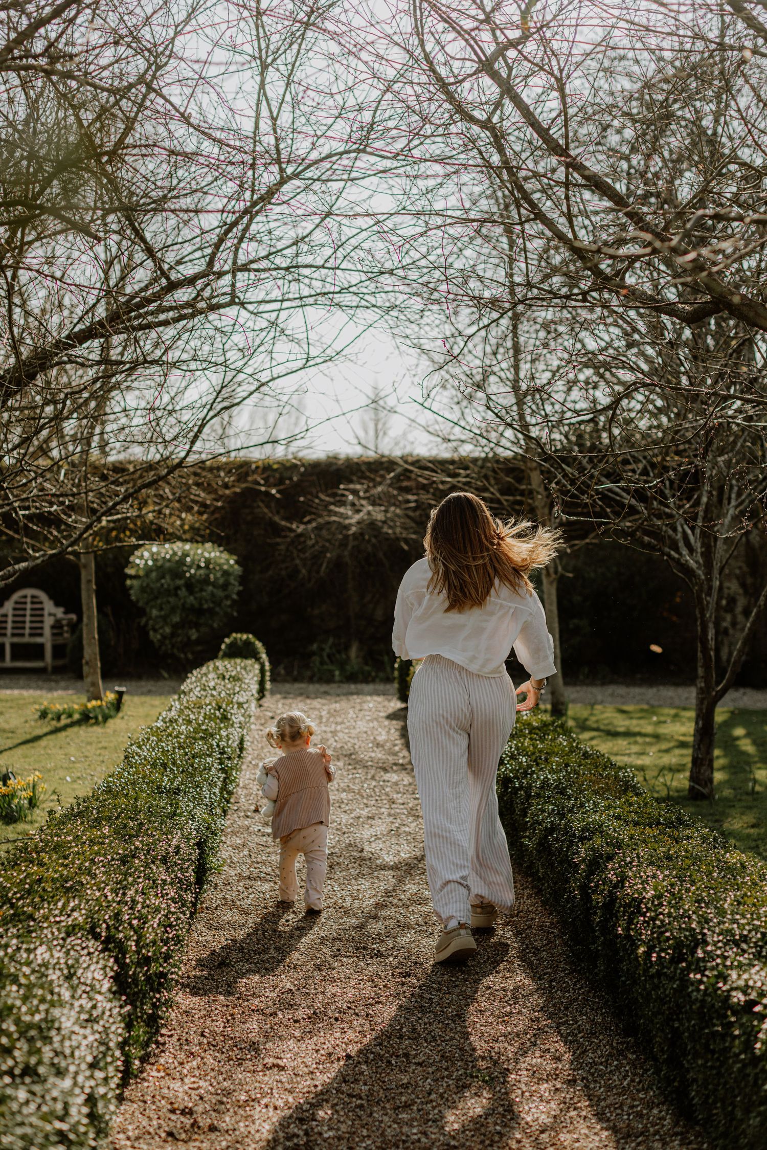Two people walking down a garden path surrounded by bare winter trees and hedges.