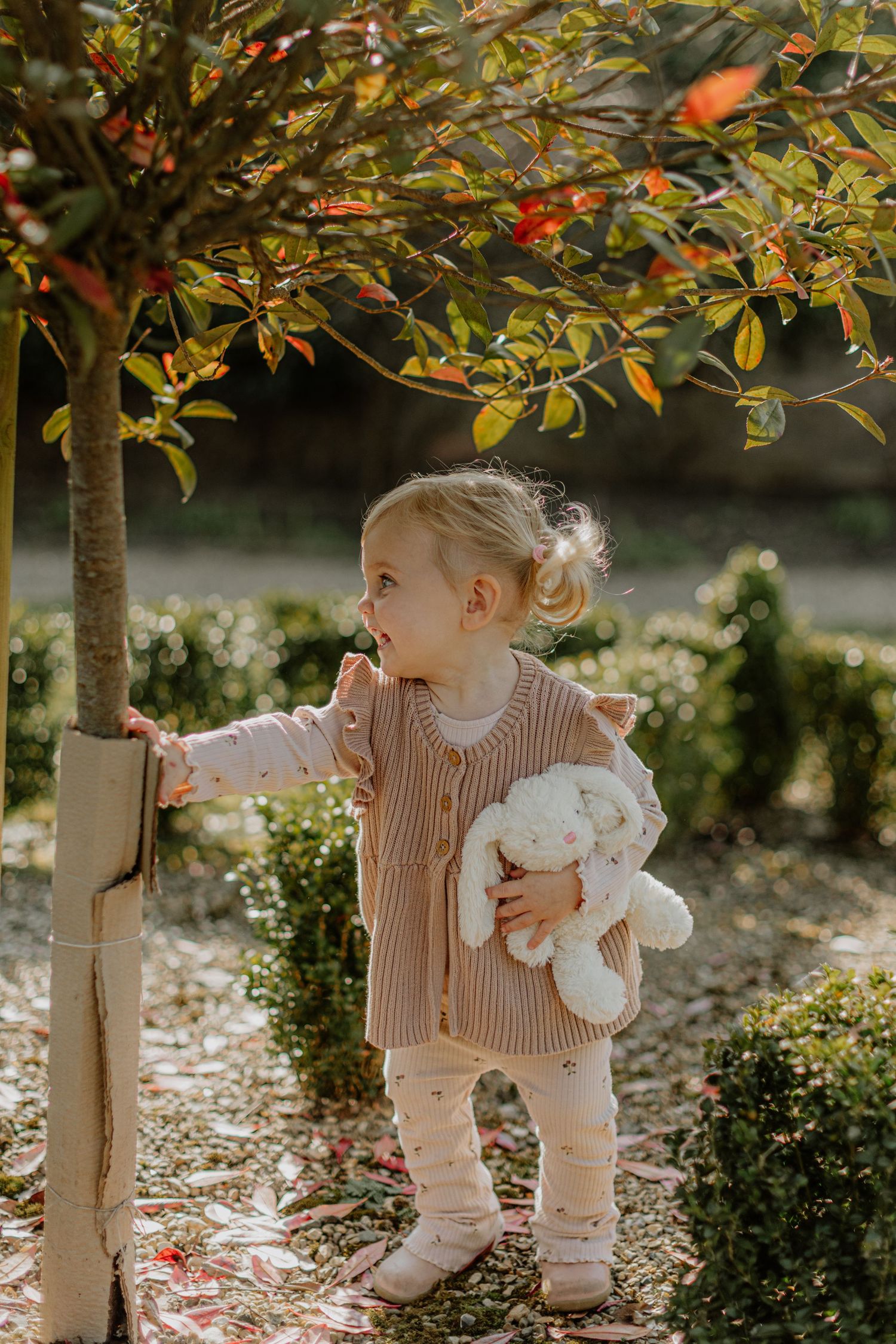 A toddler explores a garden while holding a stuffed toy beneath a fruit tree.