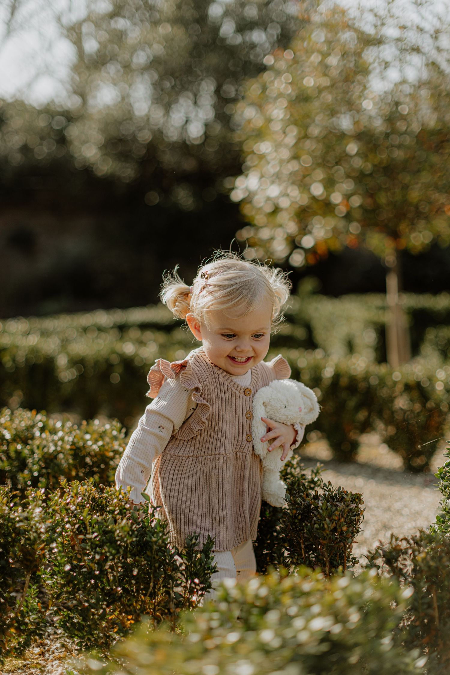 A small child smiles while playing in a garden with their stuffed animal companion.