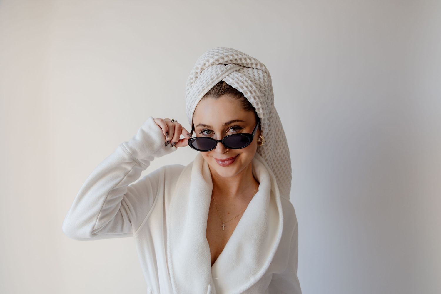 Stylish person in white robe and head wrap poses with sunglasses against white background.