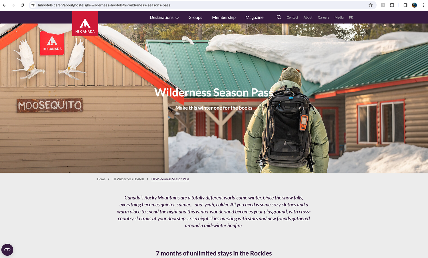 A backpacker wearing a green hiking pack stands in front of a rustic wooden wilderness lodge in snowy conditions.