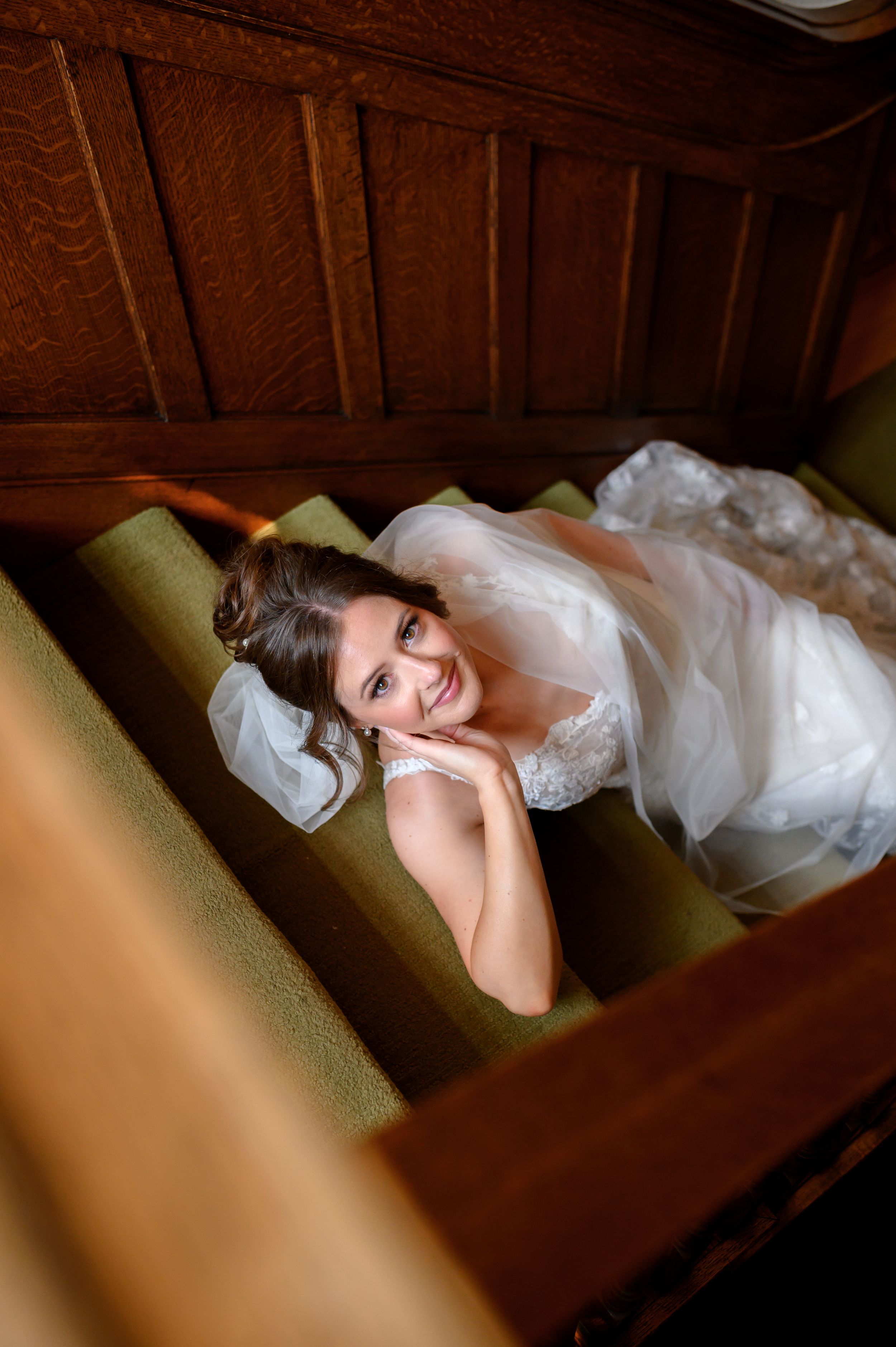 A person in a white wedding dress reclines on green upholstered stairs in a warm wood-paneled interior.