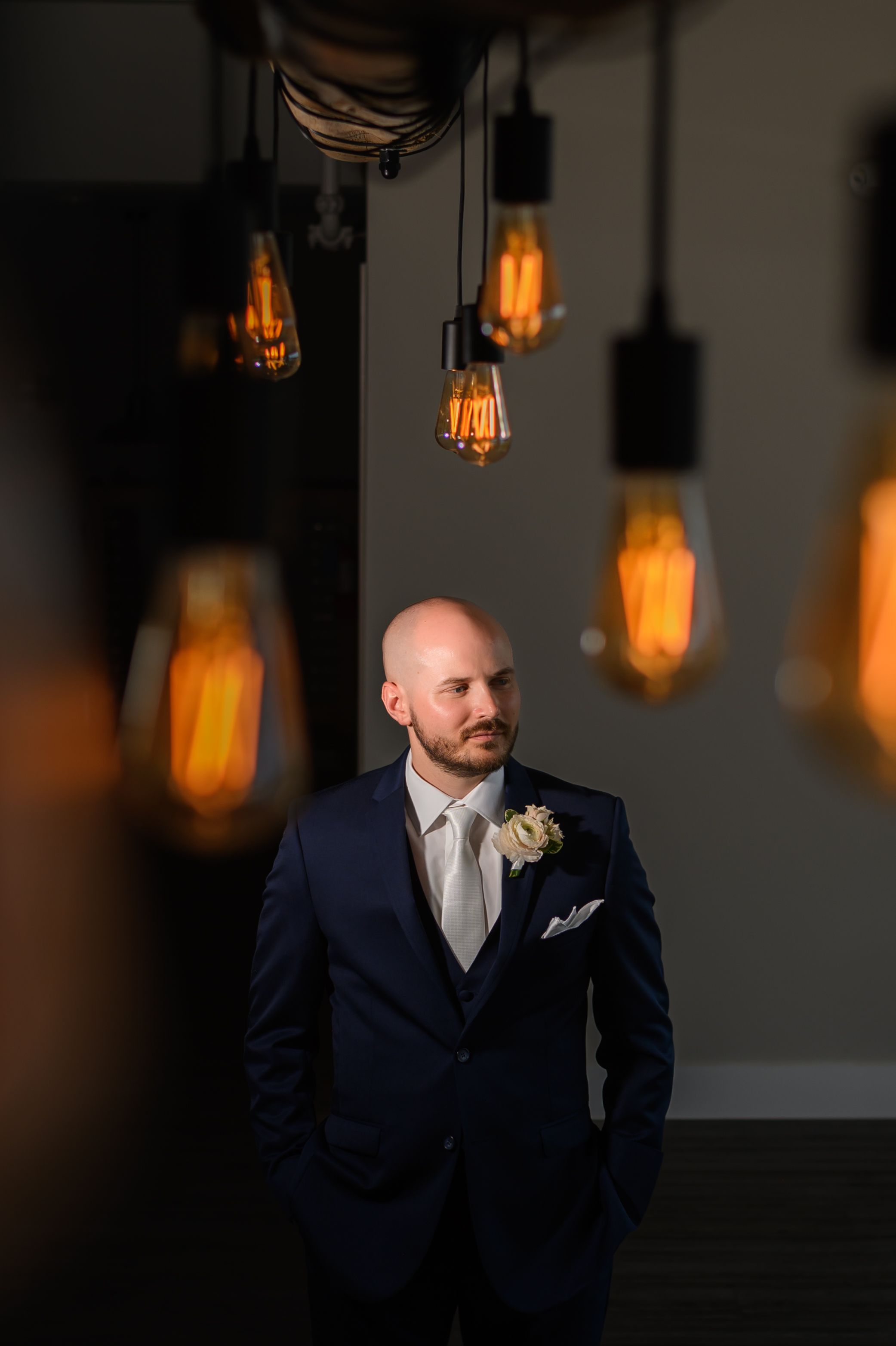 A navy blue suited figure stands in moody lighting with glowing vintage Edison bulbs hanging above in an industrial space.