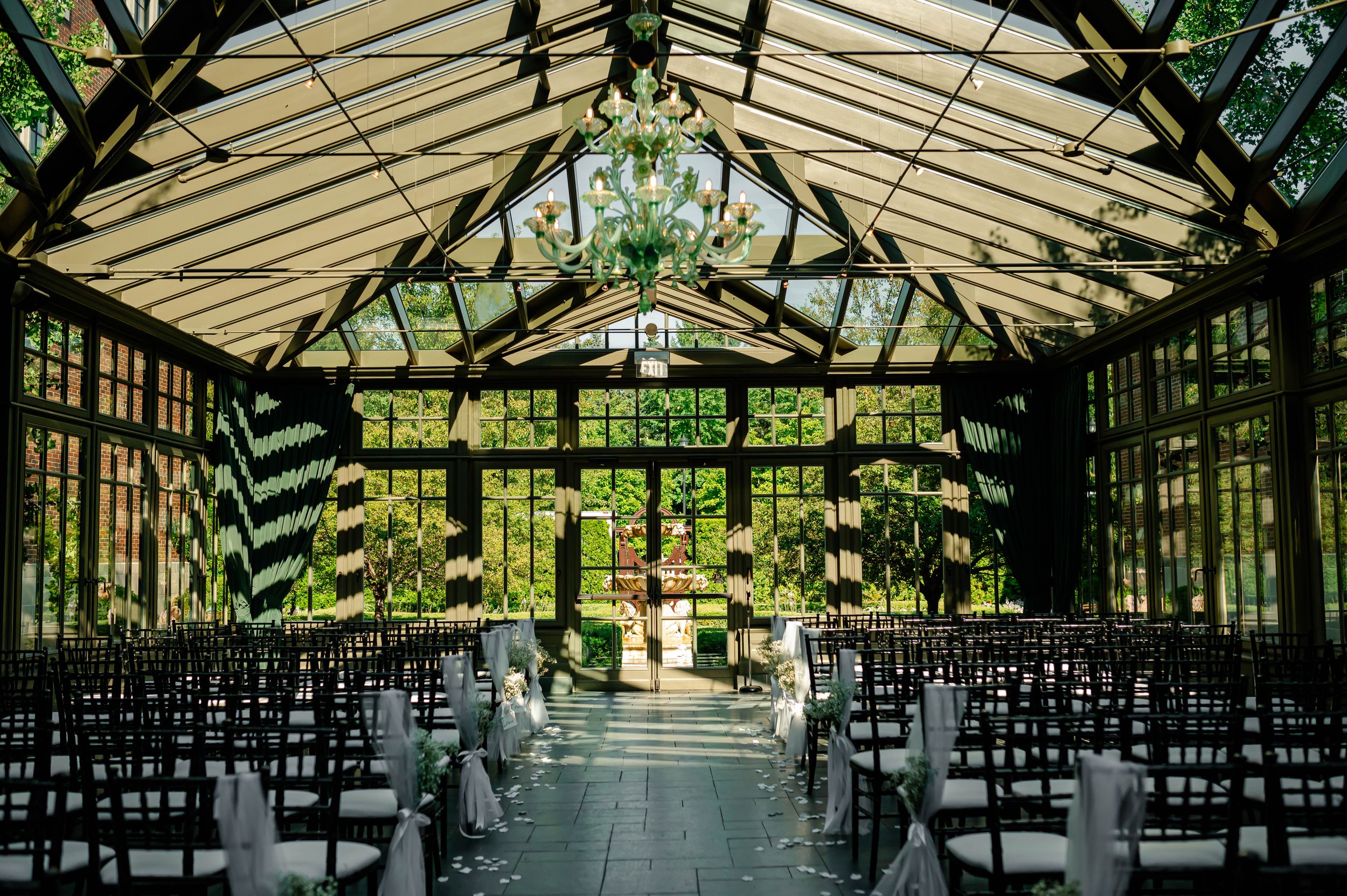 Elegant greenhouse wedding venue with rows of black chairs and natural light streaming through glass panels and greenery.