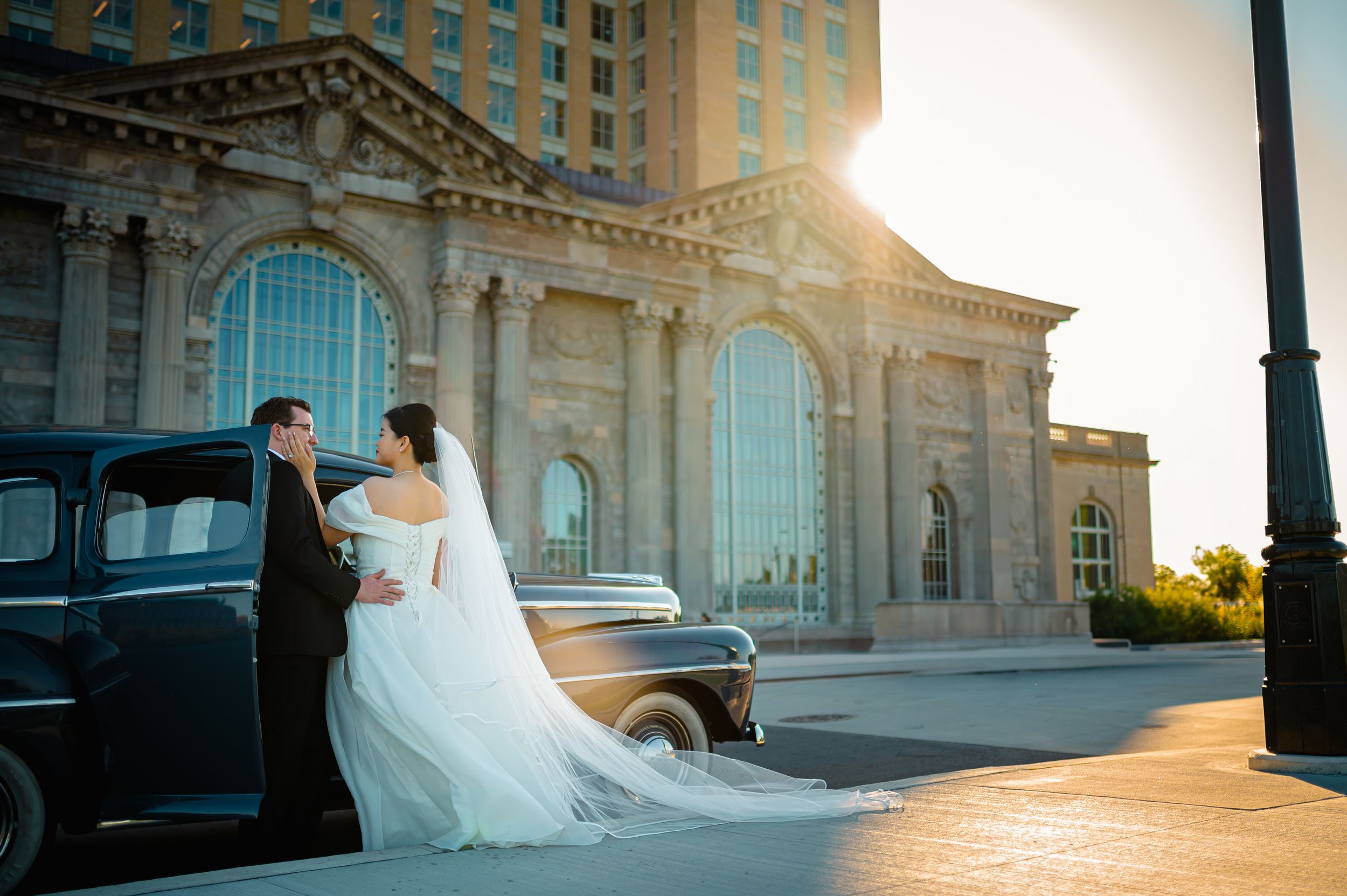 A wedding couple poses by a vintage black car at sunset in front of the Detroit Michigan Central Station