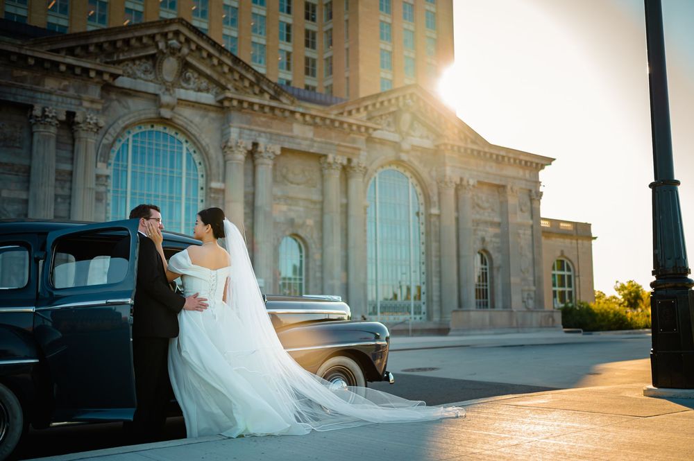 A wedding couple poses by a vintage black car at sunset in front of the Detroit Michigan Central Station