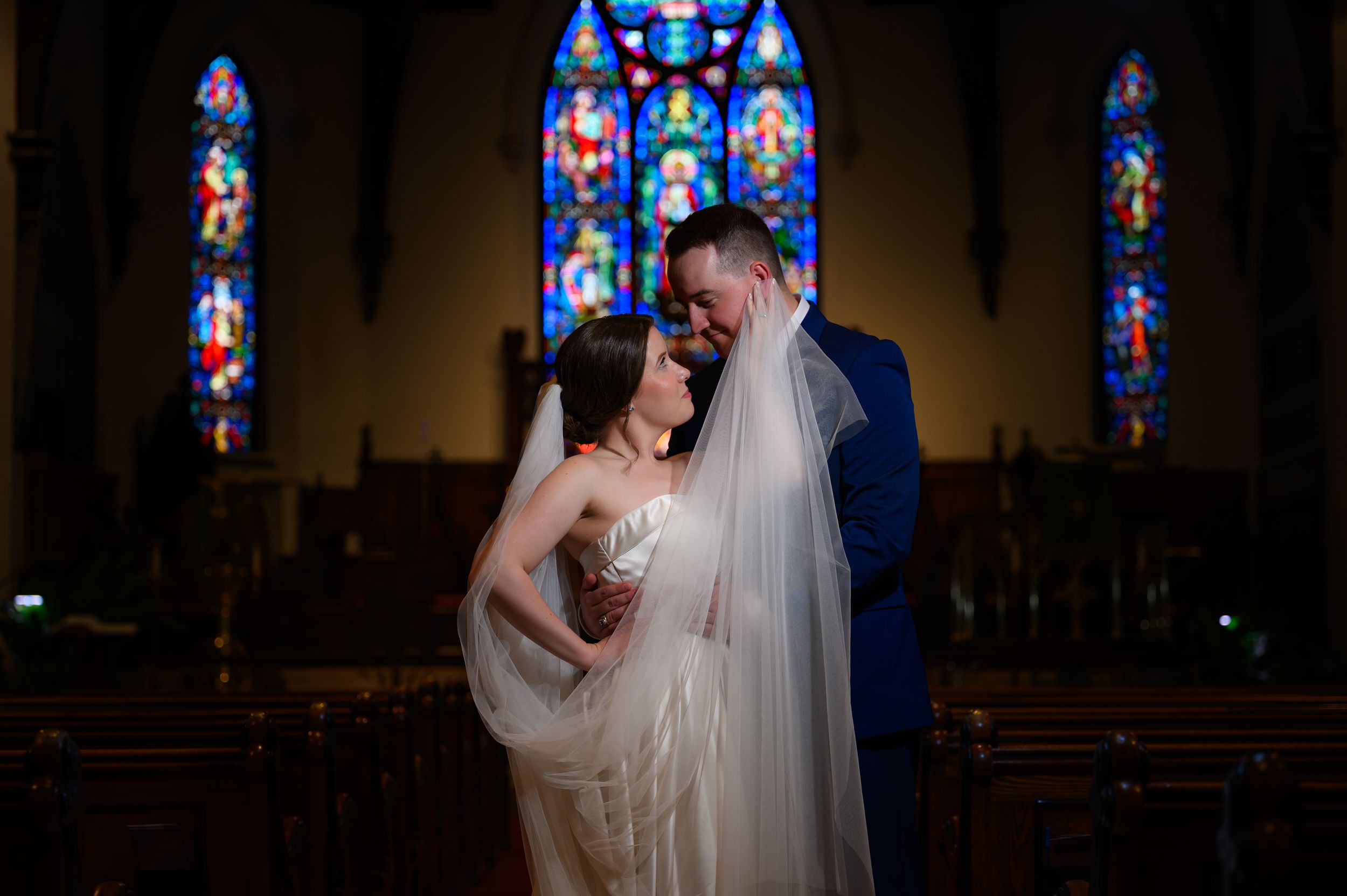 Elegant wedding couple embraces in front of stained glass windows inside a church with blue and red hues.