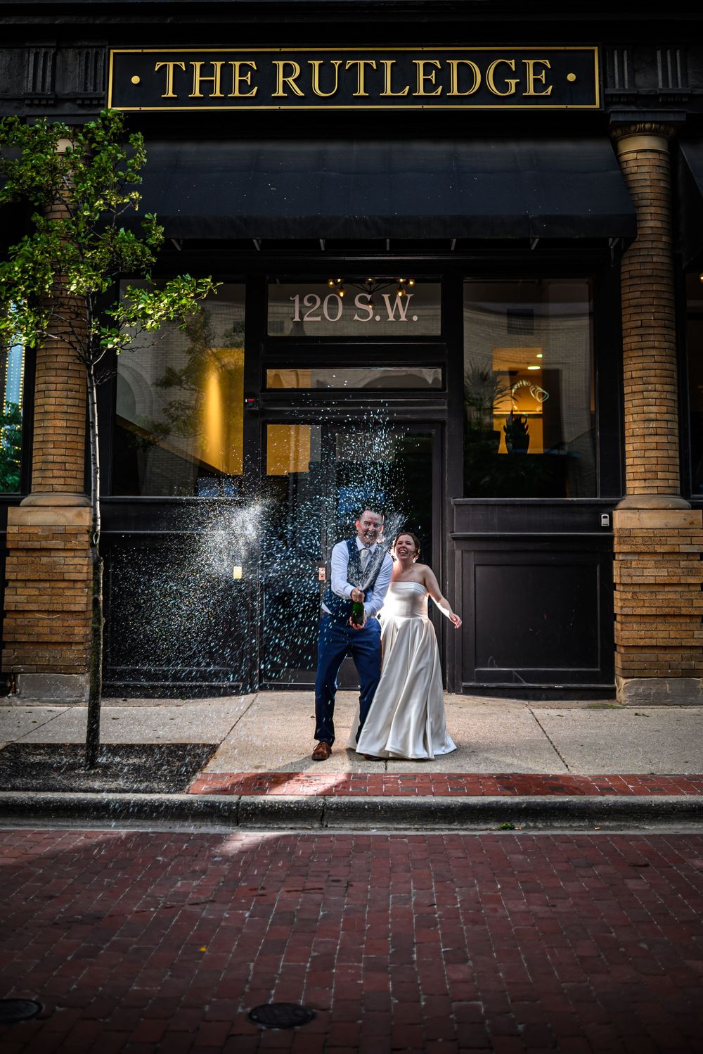 A couple stands outside The Rutledge with red brick road and dramatic splash of champagne in Grand Rapids