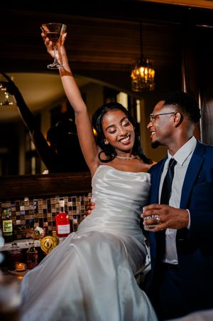 A couple in wedding attire share drinks and dance joyfully at an intimate venue with warm lighting.