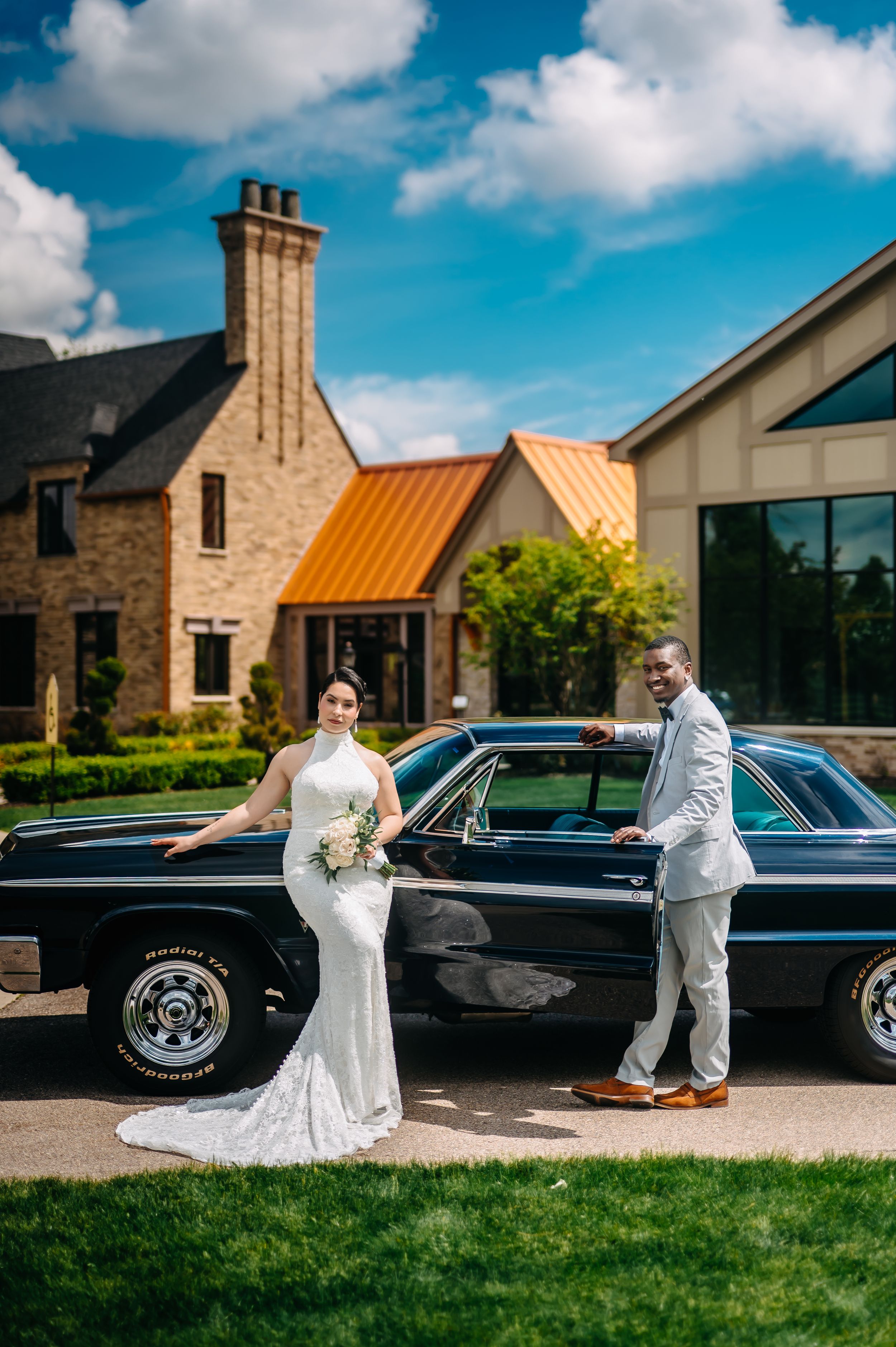 Wedding couple poses with classic vintage car outside stone building venue on sunny day with blue sky and white clouds.