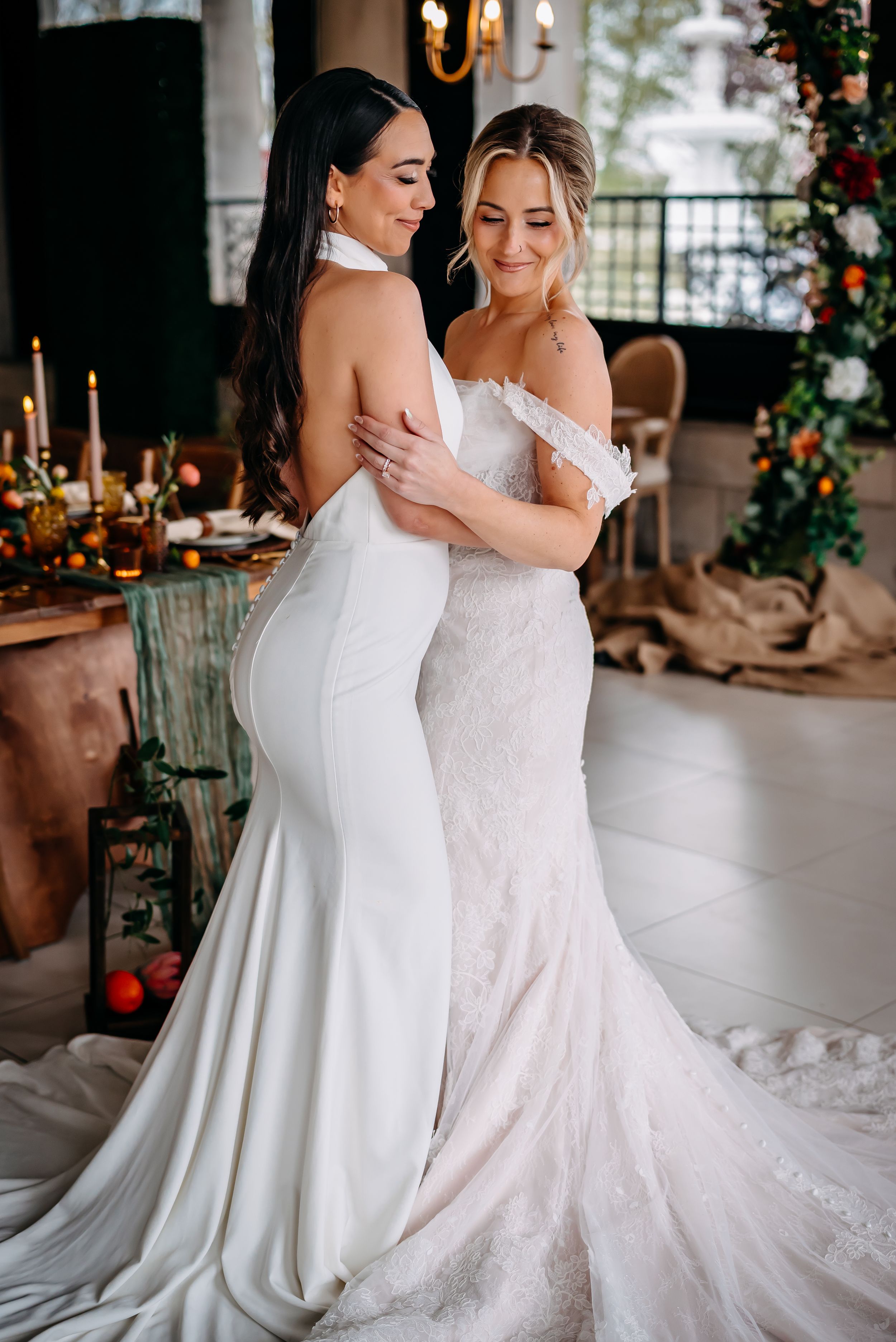 Two women in beautiful white wedding dresses embrace during an elegant indoor reception with floral decor.