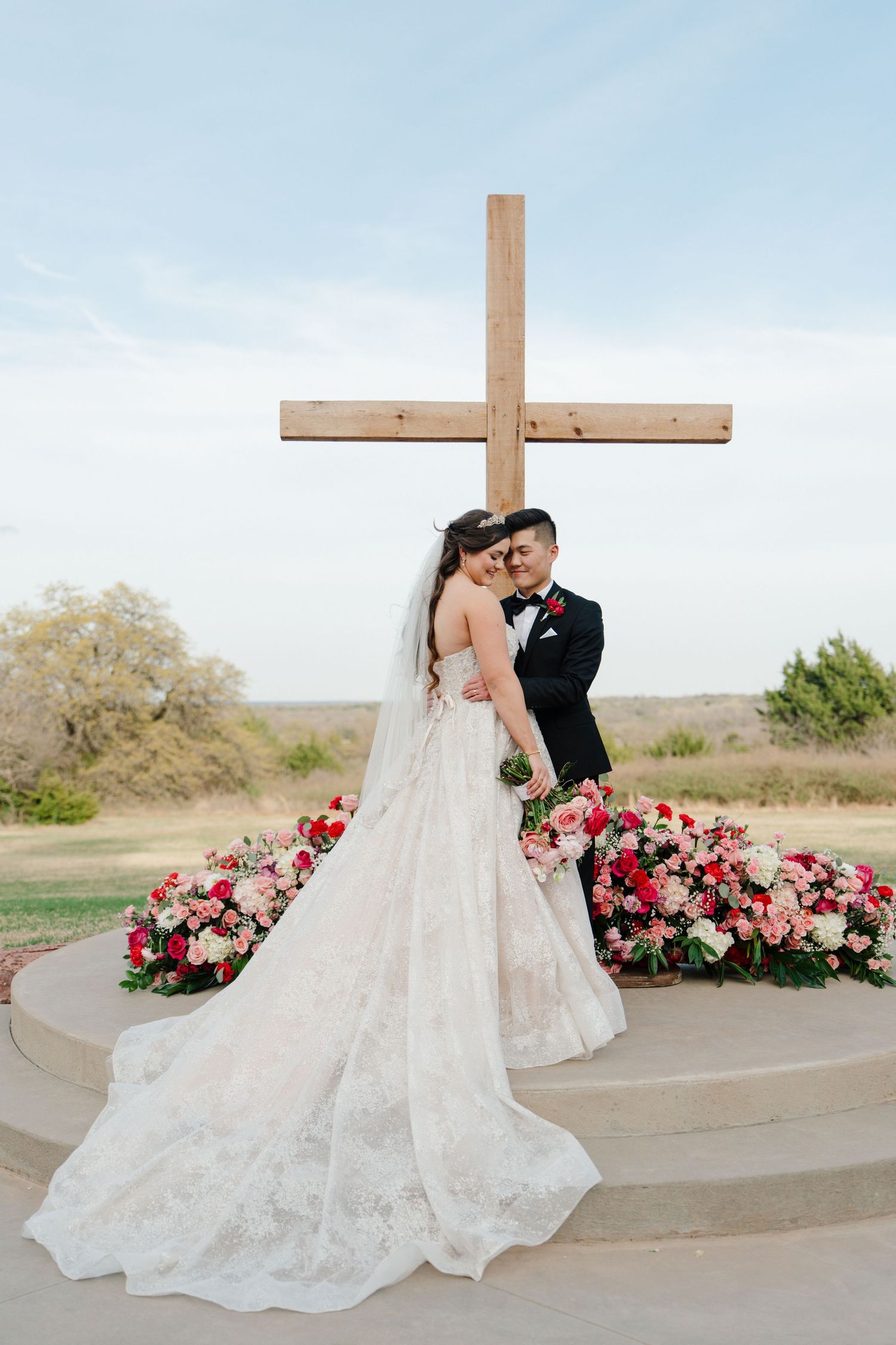 A couple stands at a wedding altar with a wooden cross and pink floral arrangements on a sunny day.