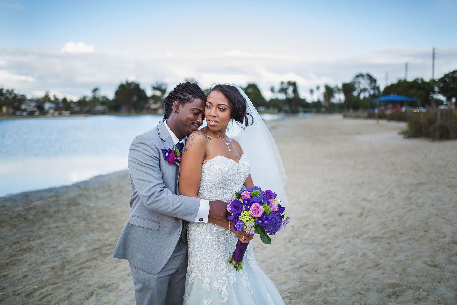 Wedding couple shares an intimate moment on a sandy beach with vibrant purple bouquet.