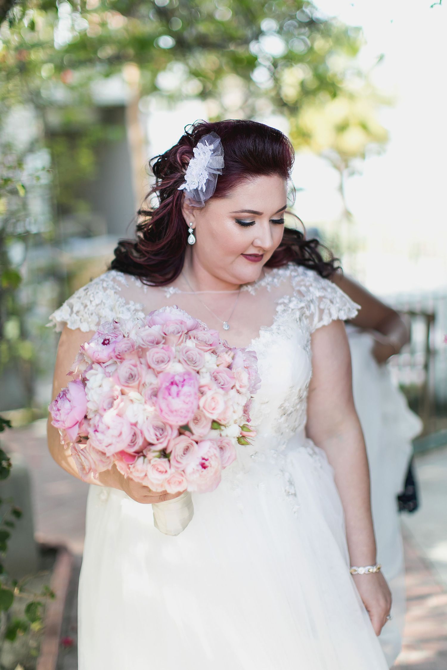 A bride in a lace dress holding a pink rose bouquet in an outdoor garden setting.