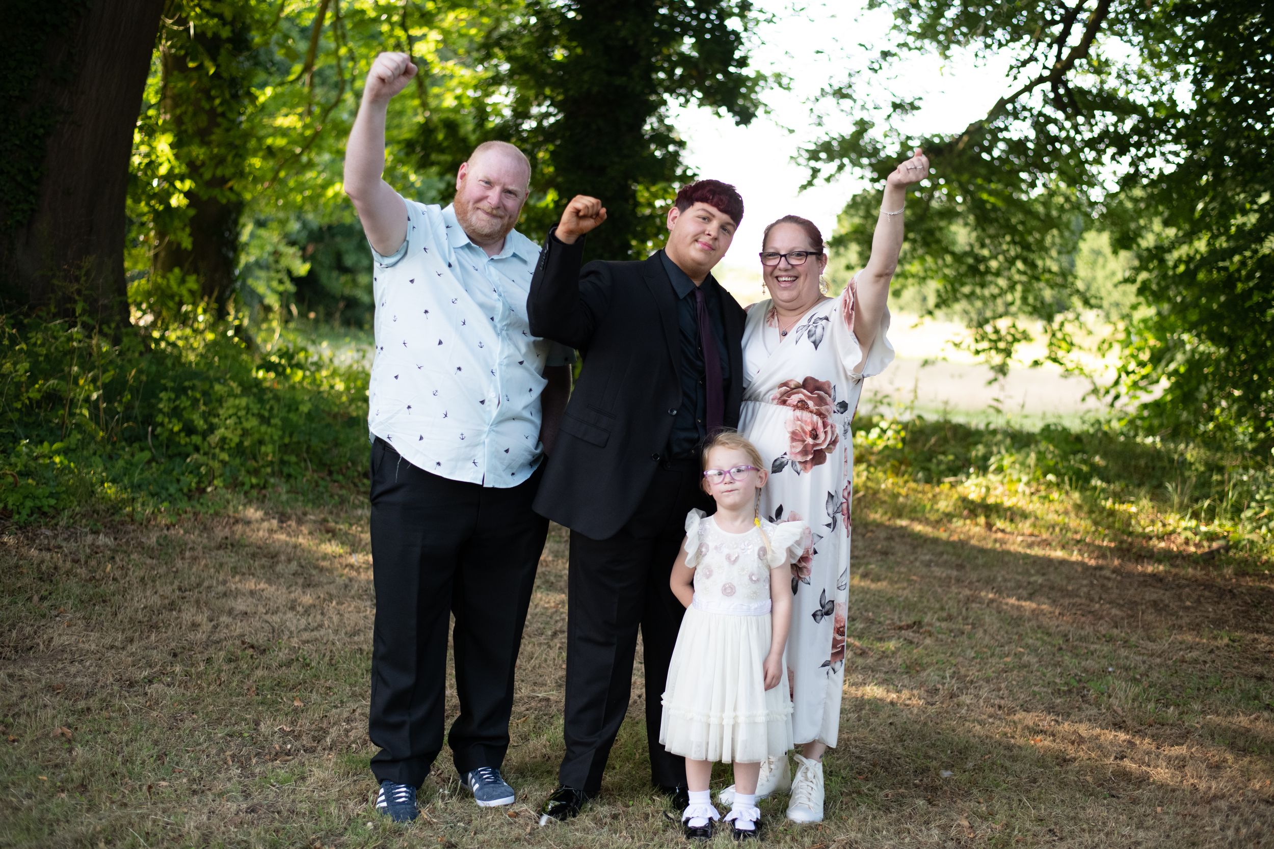 A group of five people in formal attire pose together joyfully on a sunny day in a wooded outdoor setting.