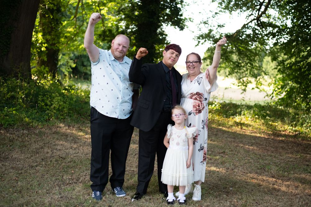 A group of five people in formal attire pose together joyfully on a sunny day in a wooded outdoor setting.