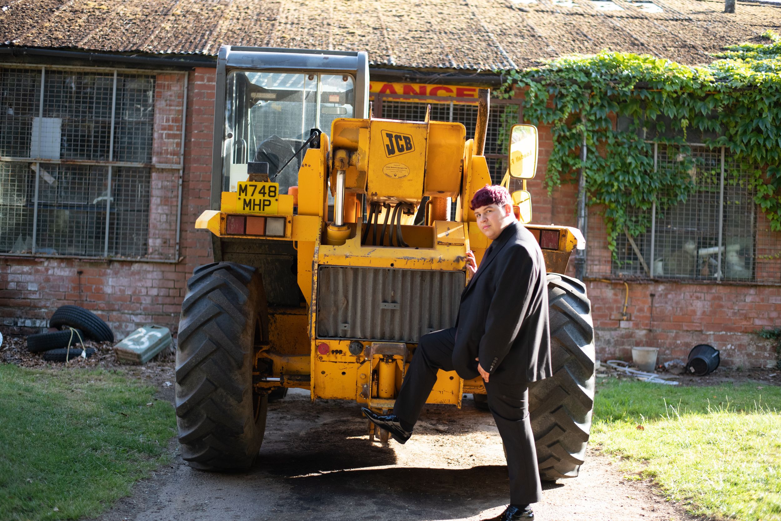 A person operates a yellow construction backhoe near a building with brick walls during daytime.