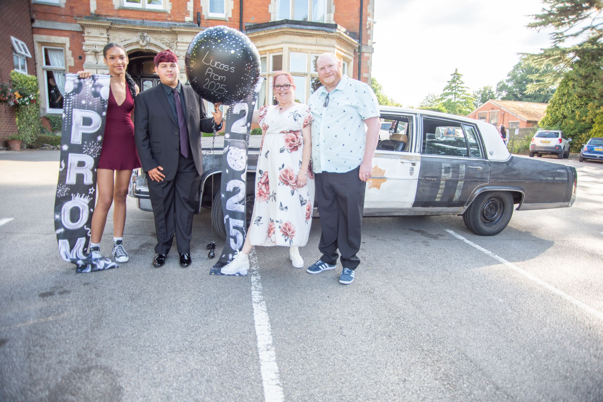 A group stands together in front of a vintage car parked outside a brick building on a sunny day.