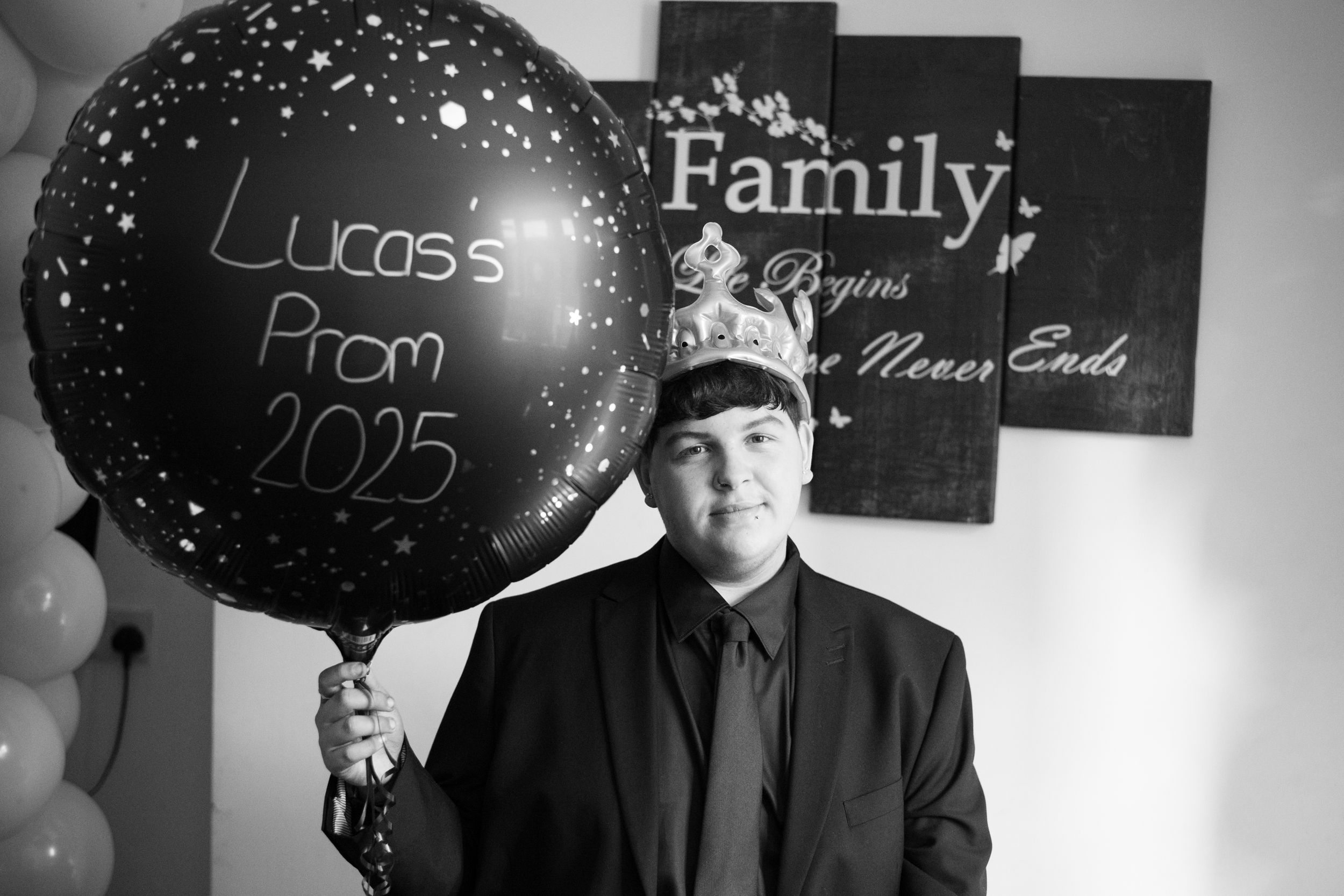 Black and white photo of someone holding a large balloon with "Lucas's Prom 2025" text against a wall with family decor.