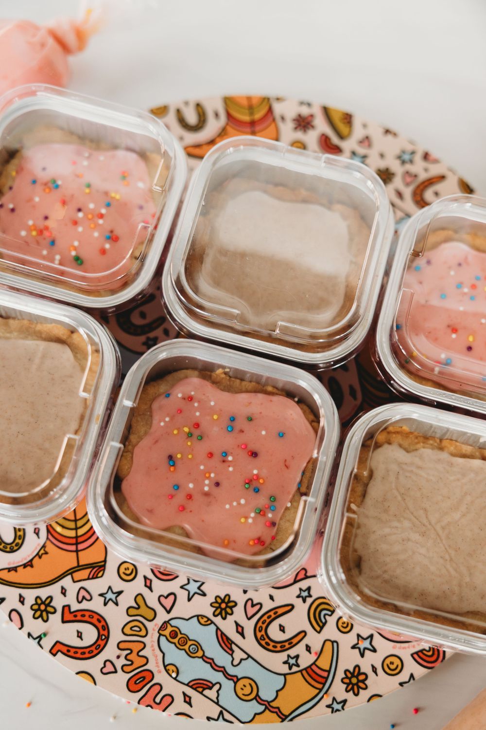Small containers of pink frosted and chocolate mini cakes decorated with colorful sprinkles on a patterned plate.