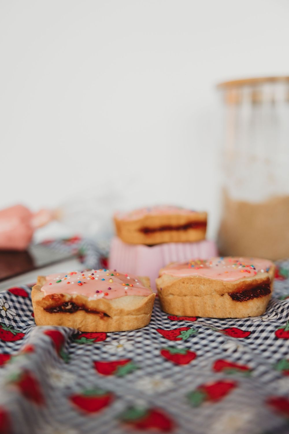 Homemade jelly-filled donuts with pink frosting and sprinkles on a red and white checkered cloth.
