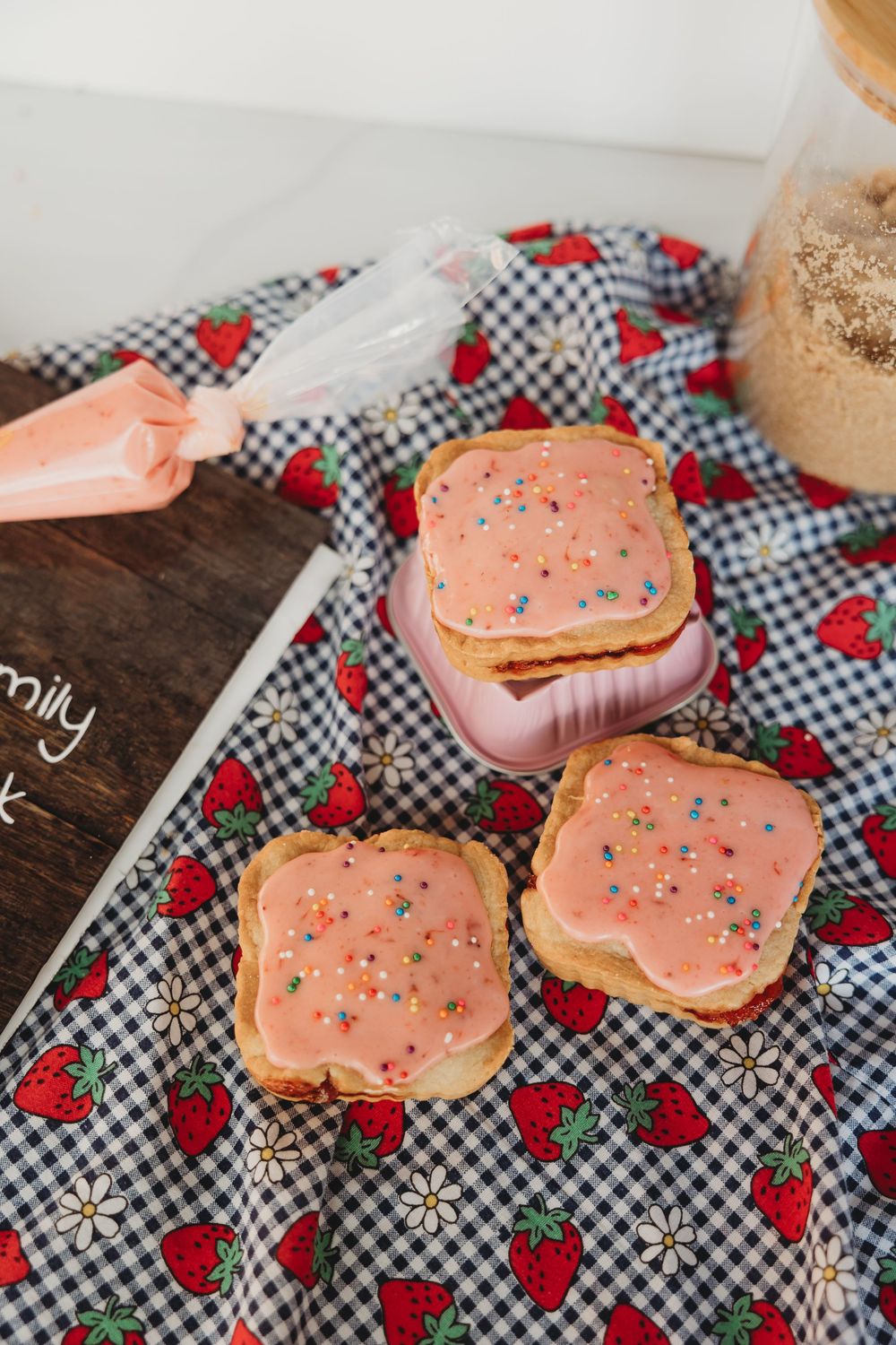 Pink frosted sugar cookies with sprinkles displayed on a red strawberry and gingham print cloth with wooden surface.