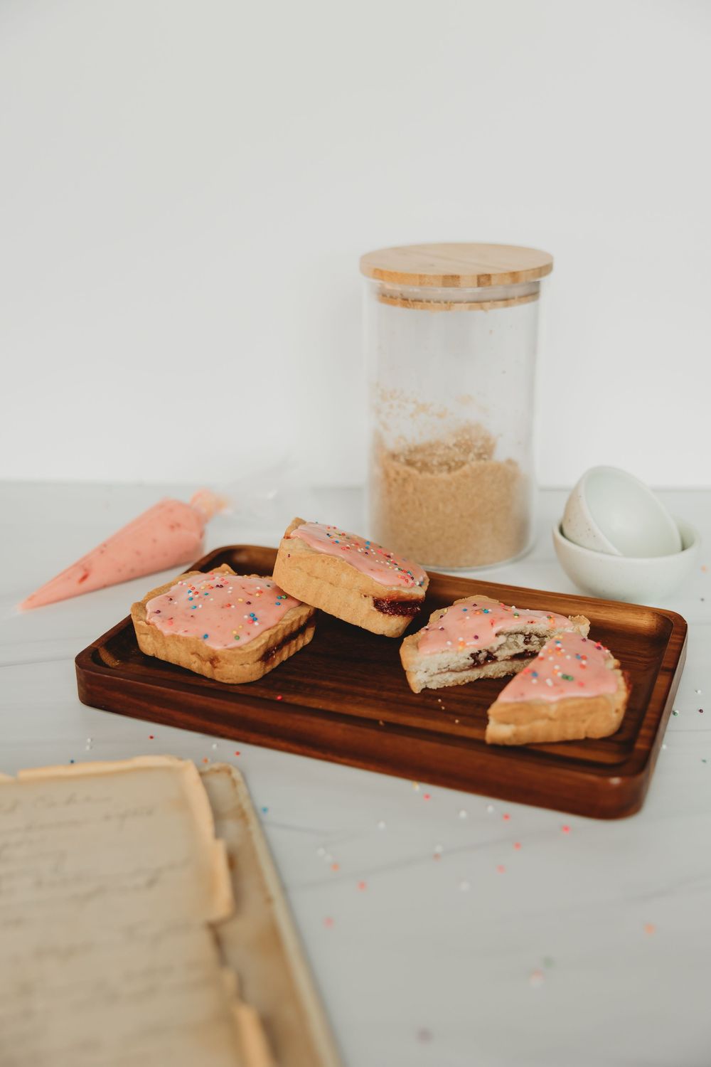 Pink frosted cookies on wooden serving board with glass jars on marble countertop.