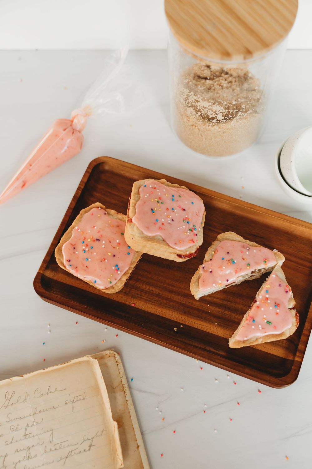 Pink frosted sugar cookies with sprinkles arranged on a wooden serving board next to baking ingredients.