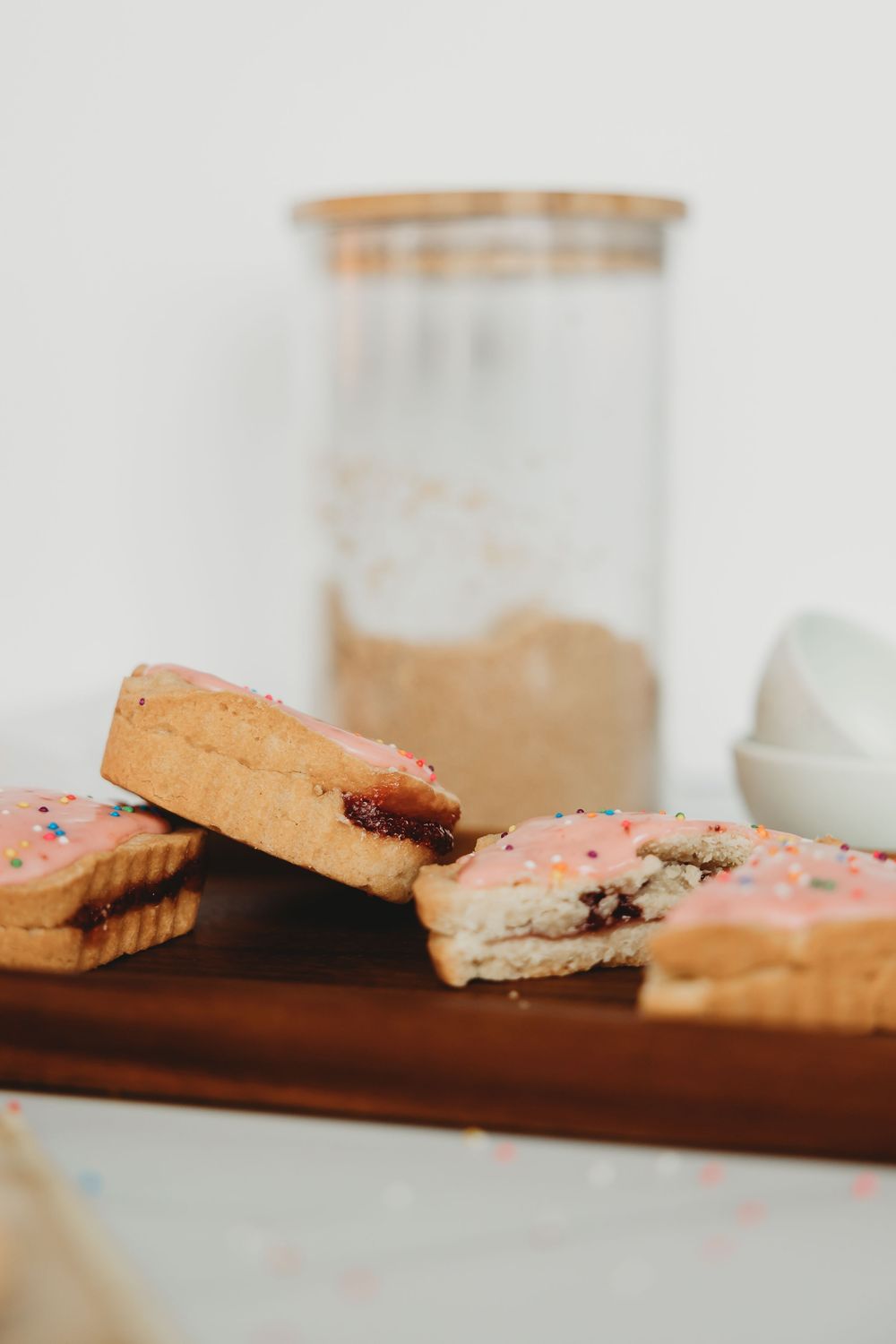 Pink-frosted sandwich cookies filled with jam displayed on a wooden cutting board next to a glass container.
