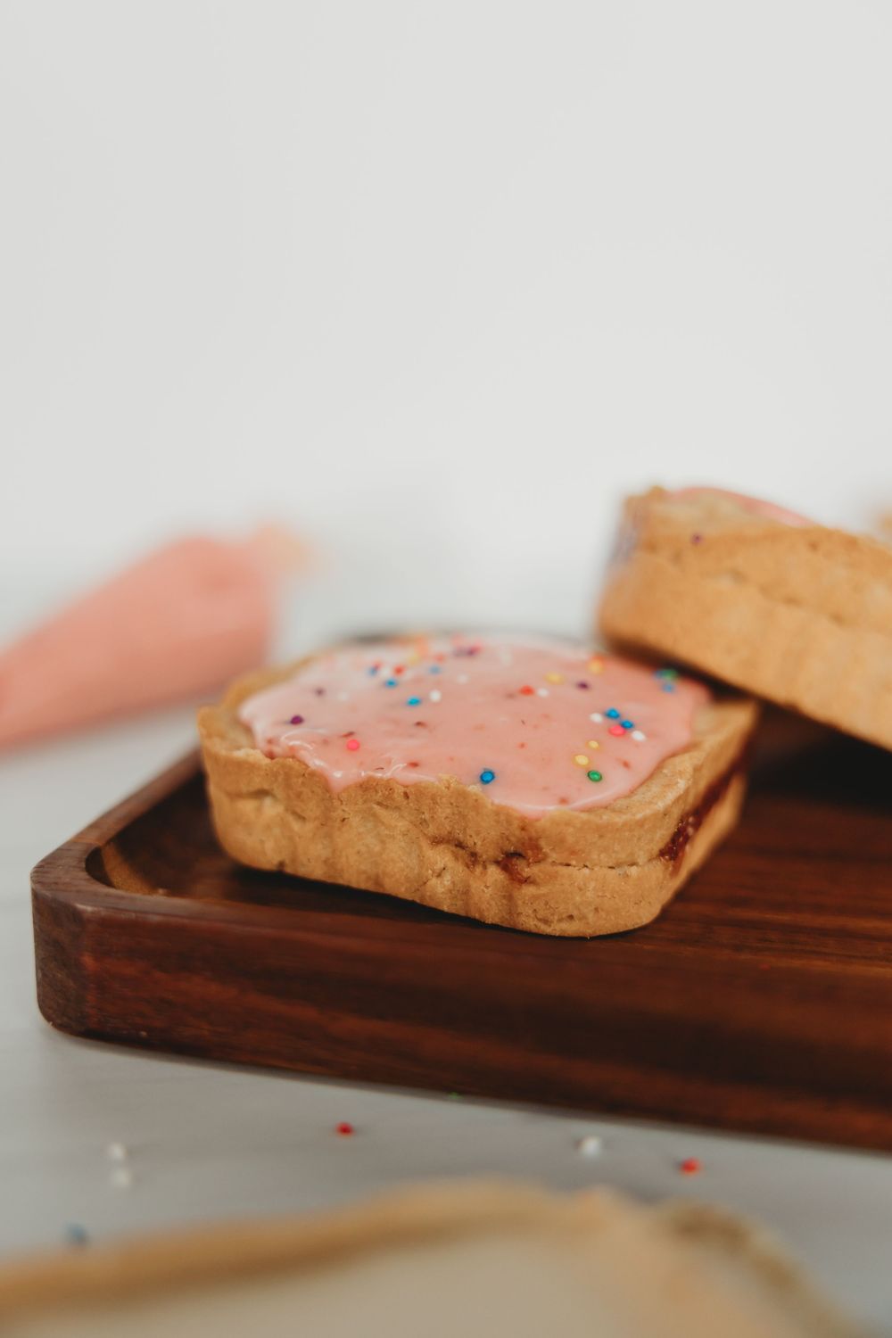 A frosted sugar cookie with pink icing and colorful sprinkles sits on a wooden serving board.