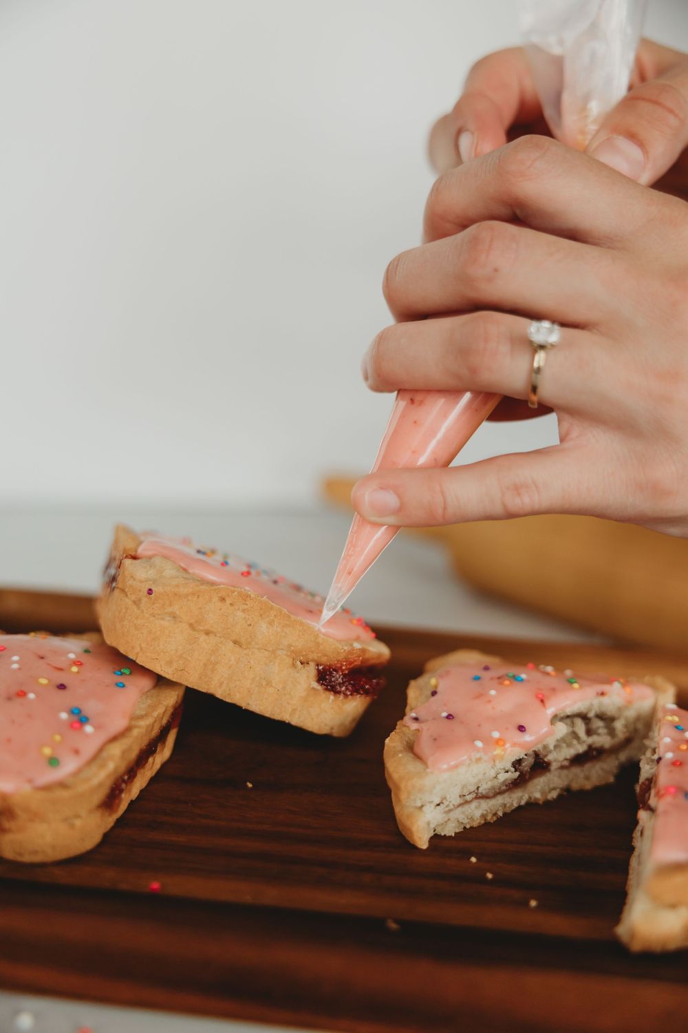 Hands with an engagement ring pipe pink frosting onto homemade cookies on a wooden cutting board.