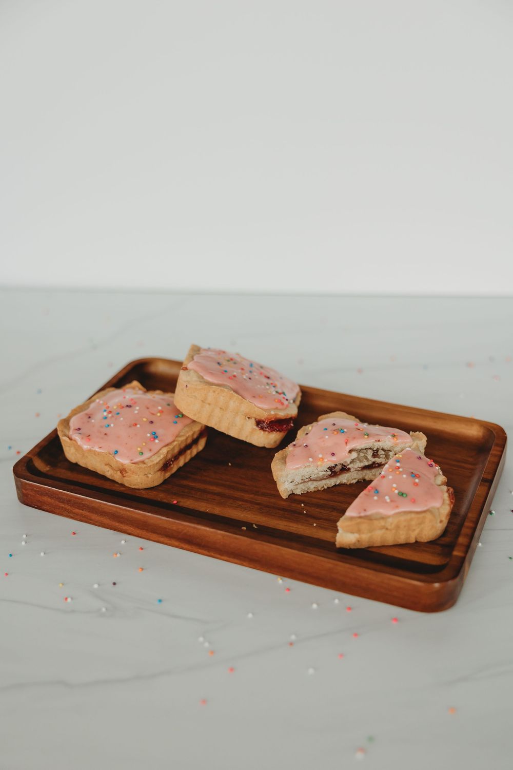 Pink frosted sugar cookies with rainbow sprinkles arranged on a rustic wooden serving board in diagonal line.