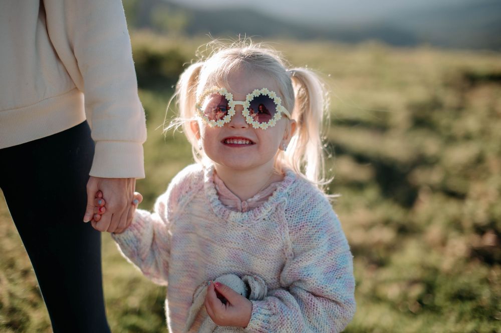Child wearing sunglasses and pink sweater smiles while holding someone's hand outdoors on sunny day.
