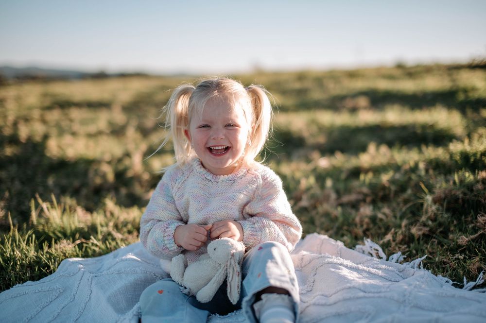 A baby wearing a white sweater sits and plays on a blanket in a grassy field at sunset.