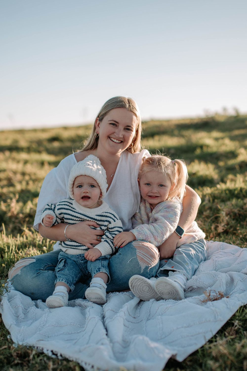 A family sits together on a white blanket in a grassy field during golden hour while sharing tender moments together.
