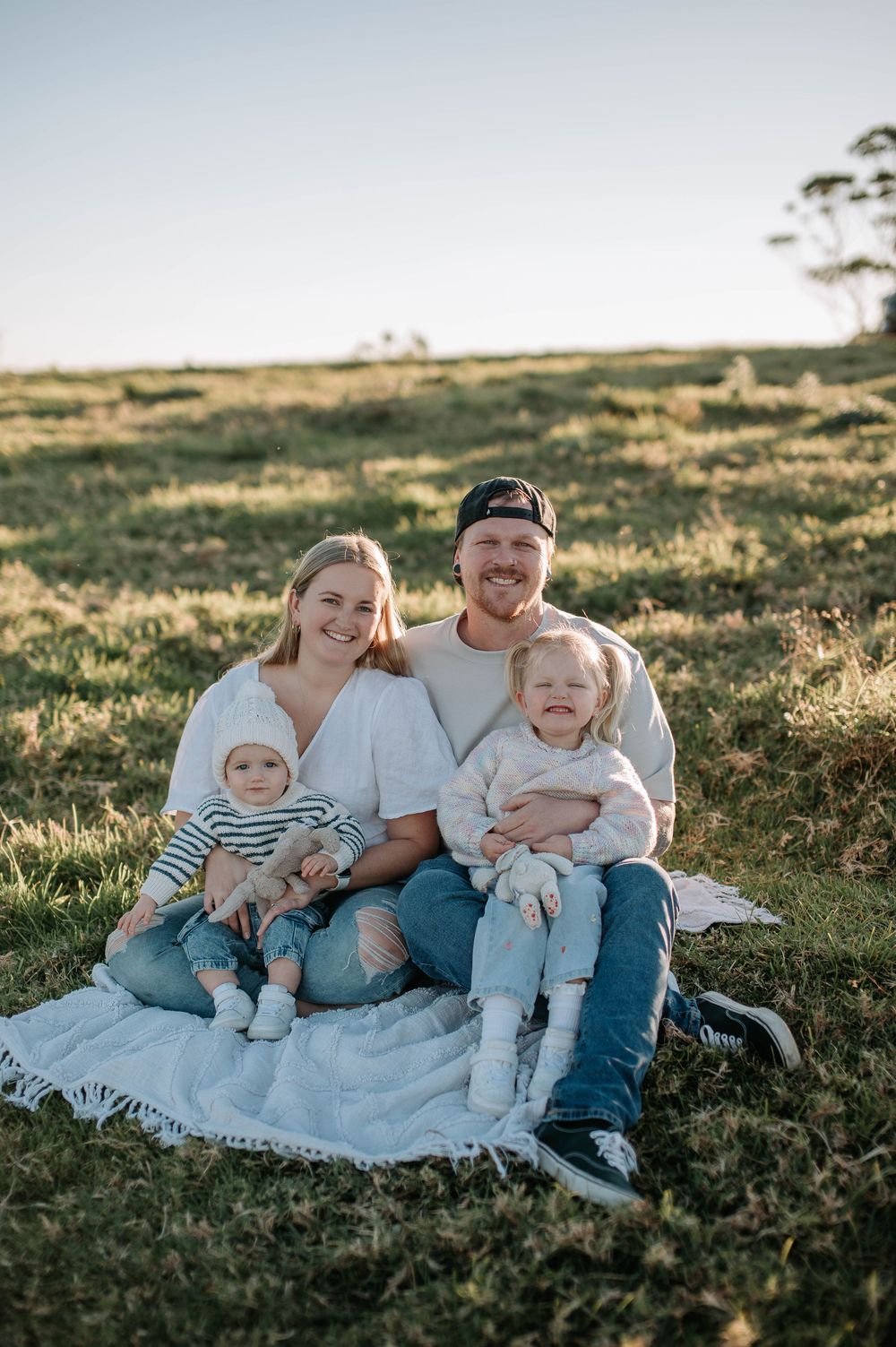 Family sitting together on white blanket in grassy field during golden hour sunset.