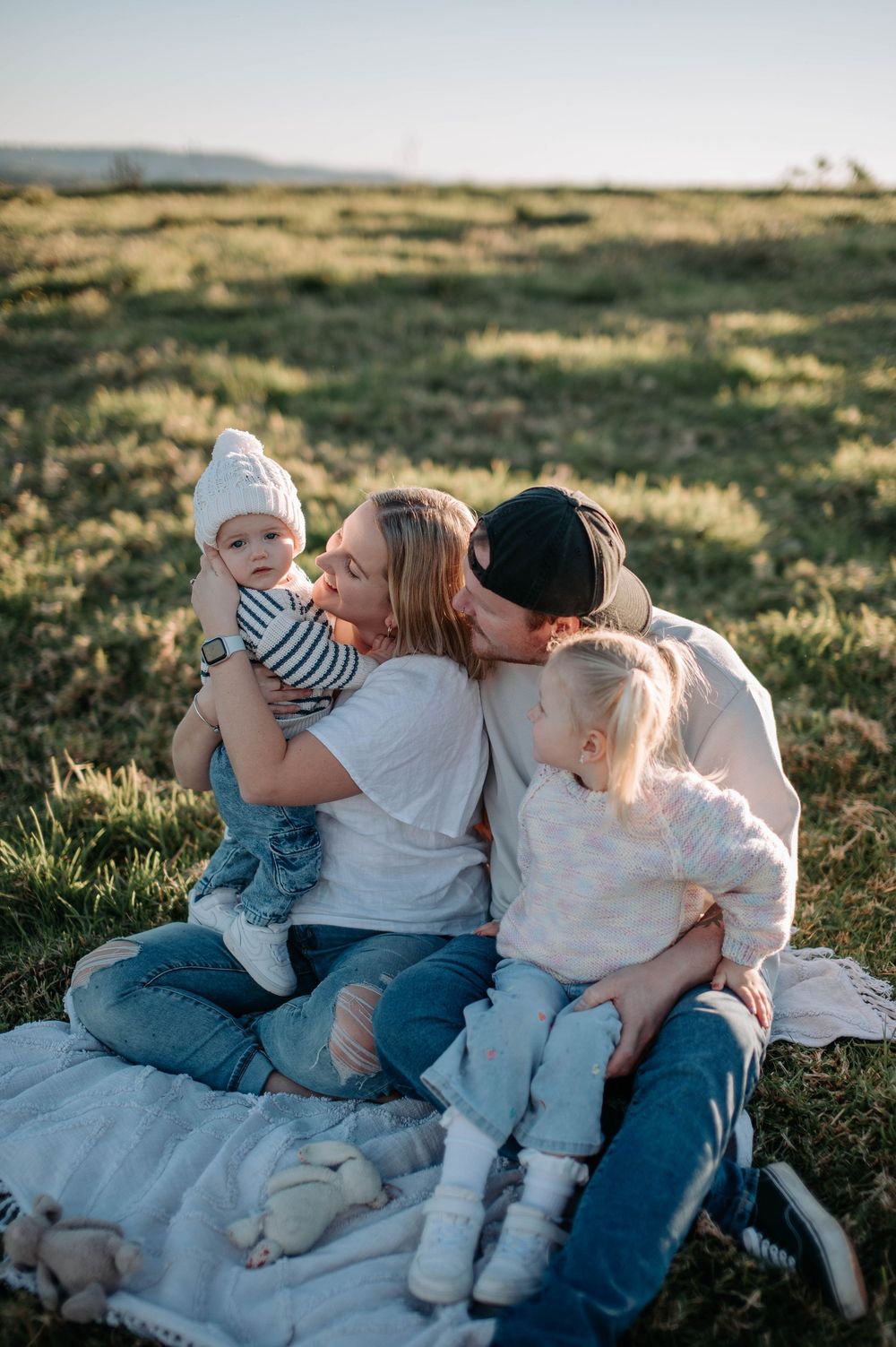 A family sits on a blanket in a grassy field during golden hour, sharing an intimate moment together outdoors.