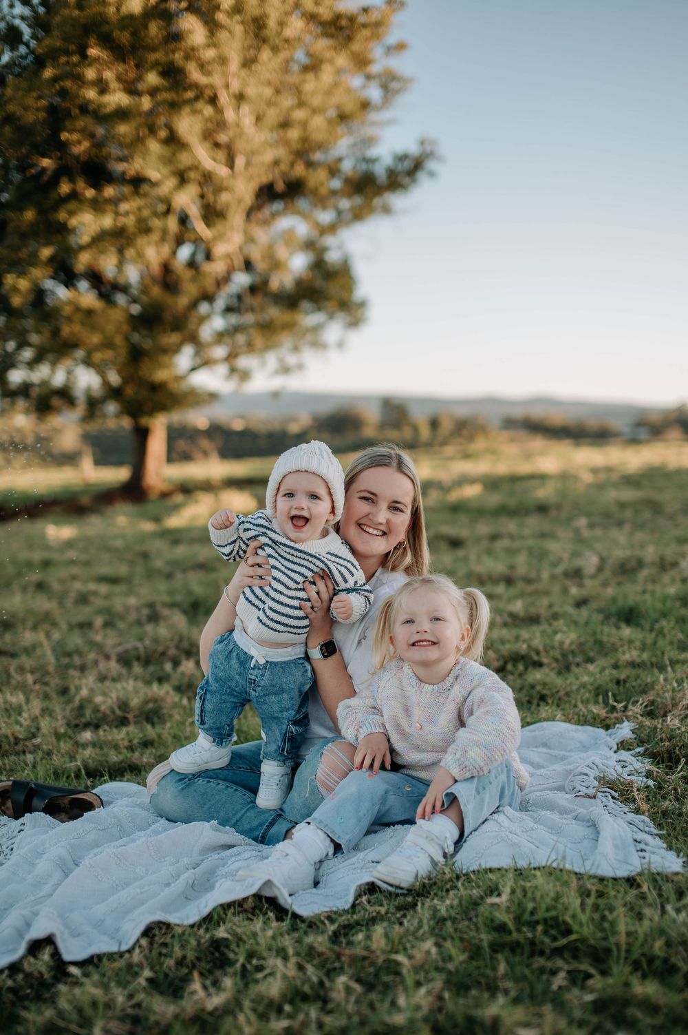 Family sits together on white blanket during golden sunset in grassy field with trees in background.