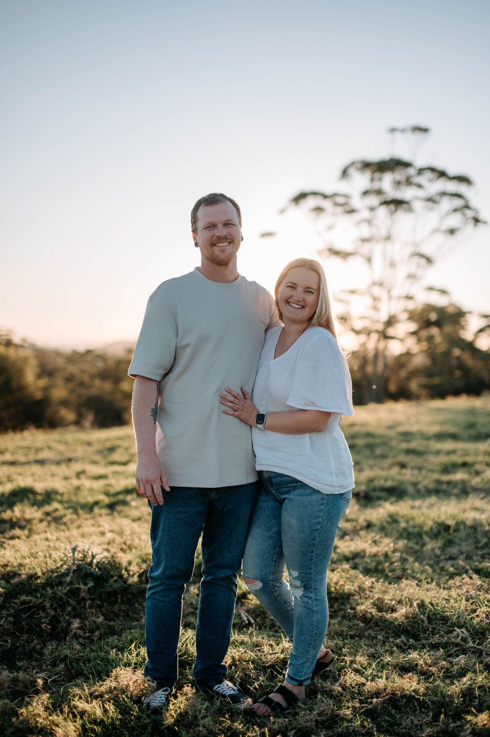 A couple embraces in a sunlit meadow during golden hour wearing casual white shirts and jeans.
