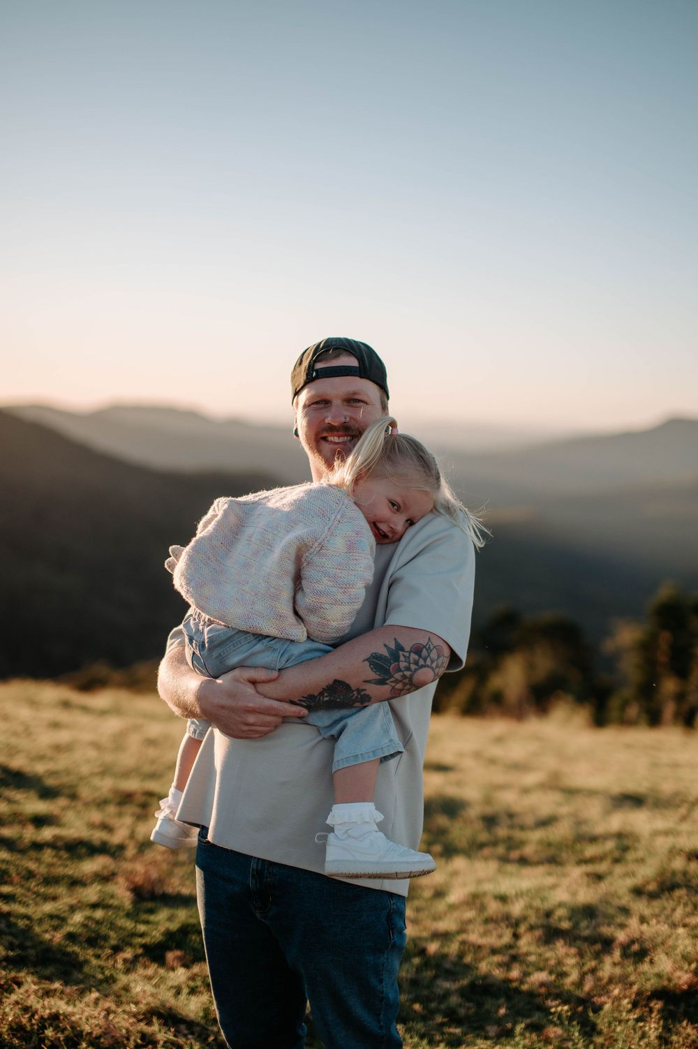 Two people sharing an affectionate embrace in a sunlit meadow with mountains in the golden hour background.