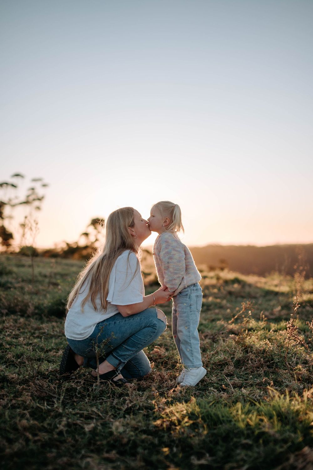 A mother and child share a tender moment in a grassy field at sunset.