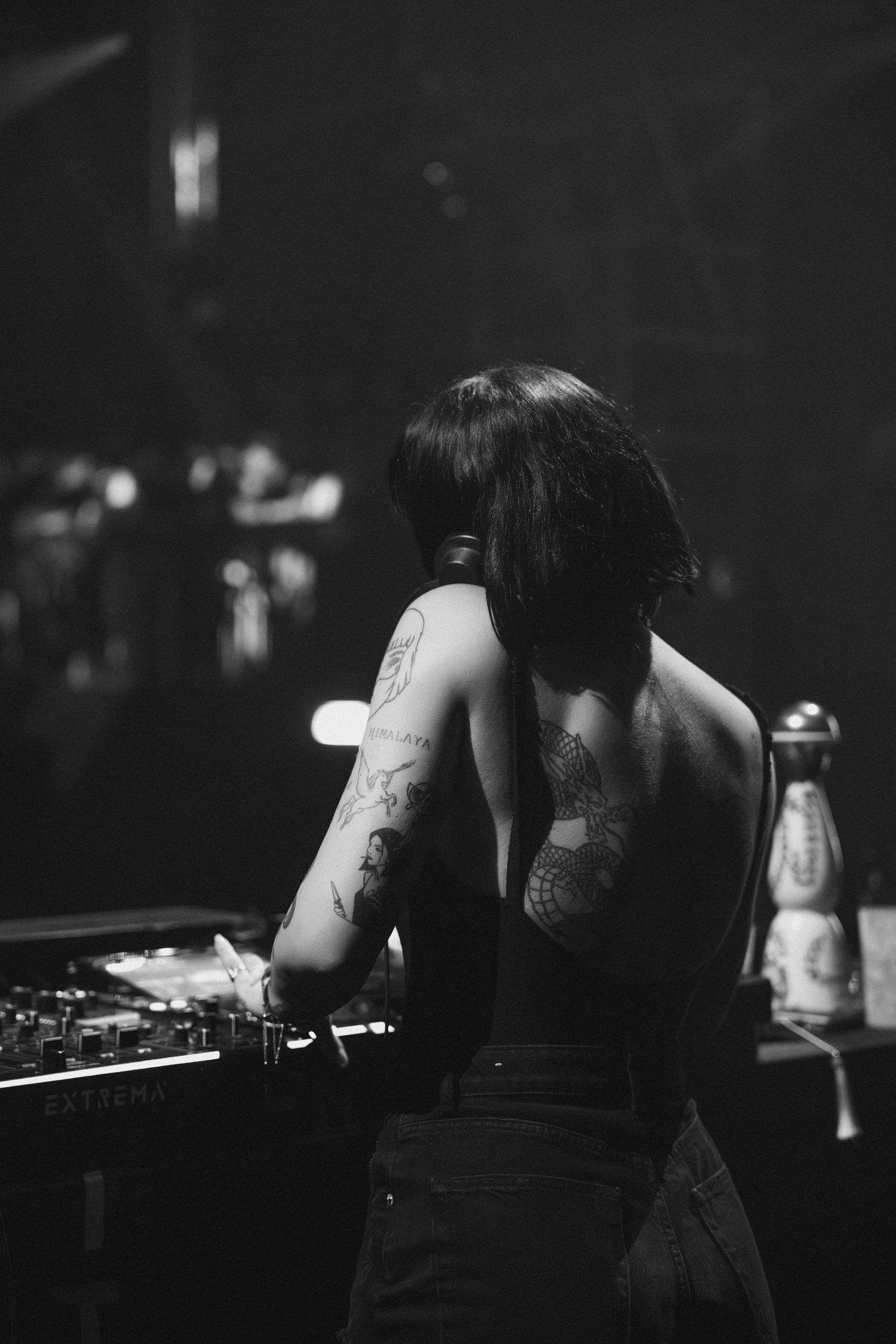 Black and white photo of a DJ performing at a club showing tattoos and dark hair from behind.