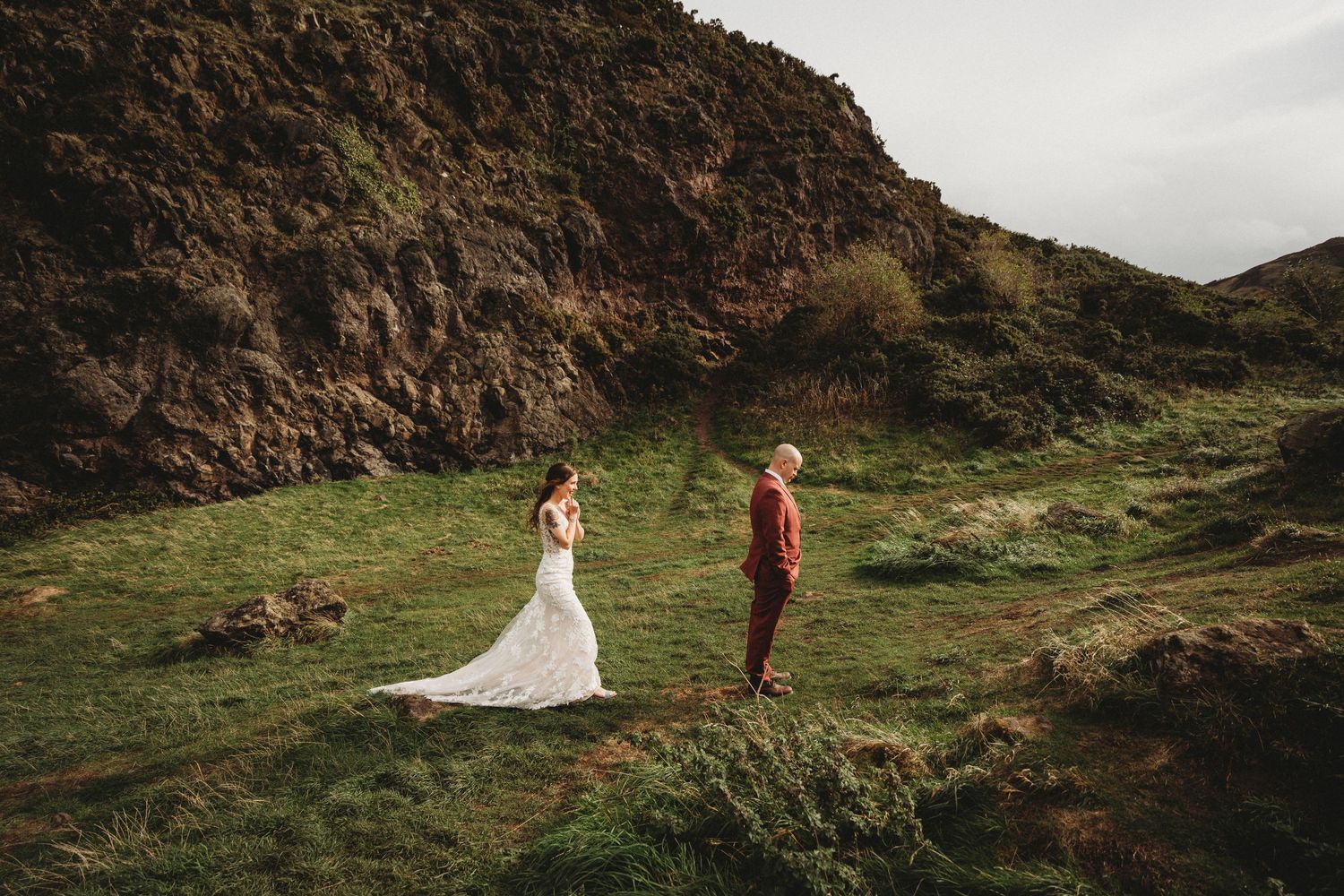 Bride and groom stand apart on a dramatic hillside during their wedding photoshoot.
