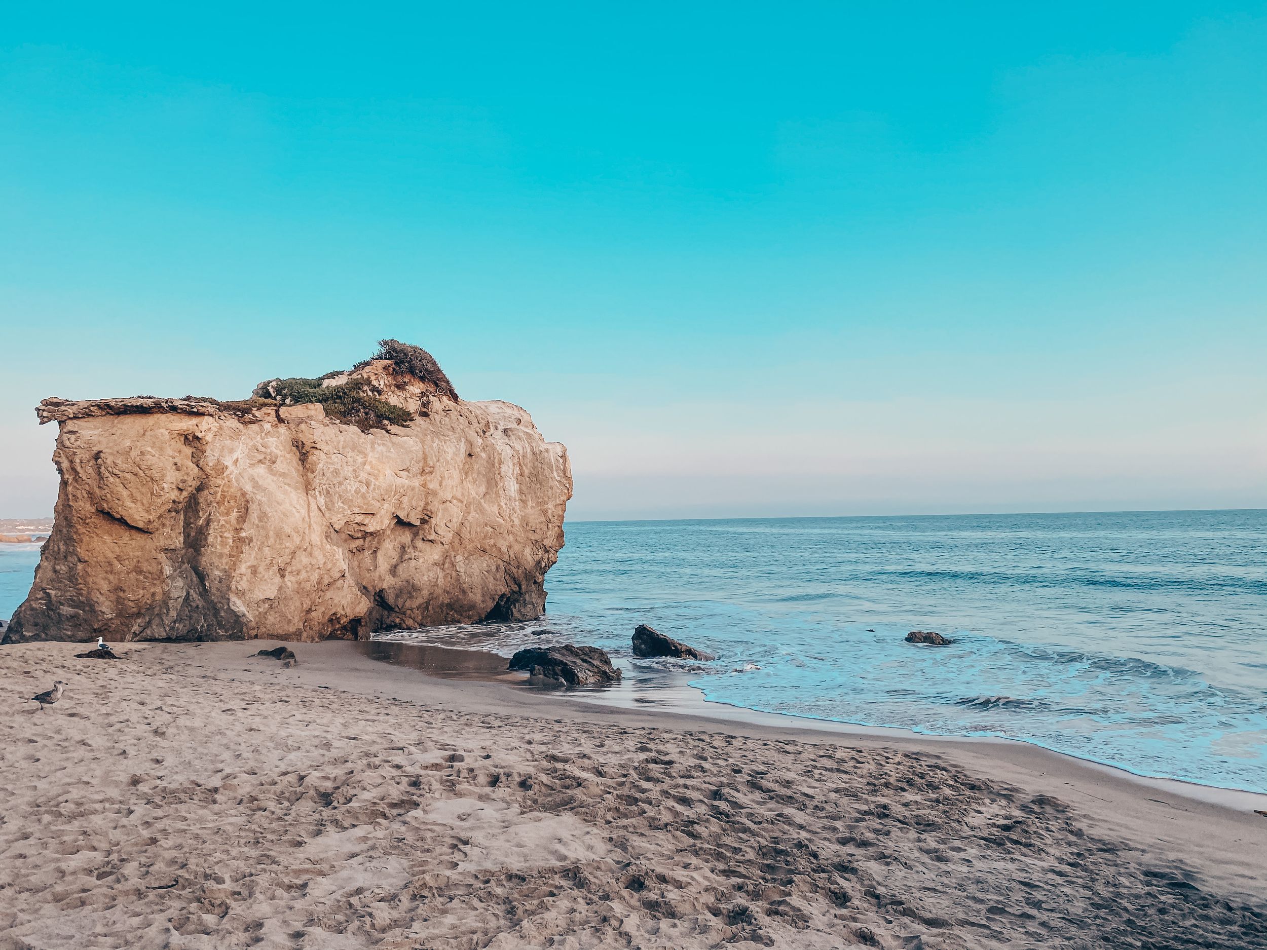 Scenic coastal view of a large limestone rock formation on a sandy beach against turquoise waters and clear blue sky.